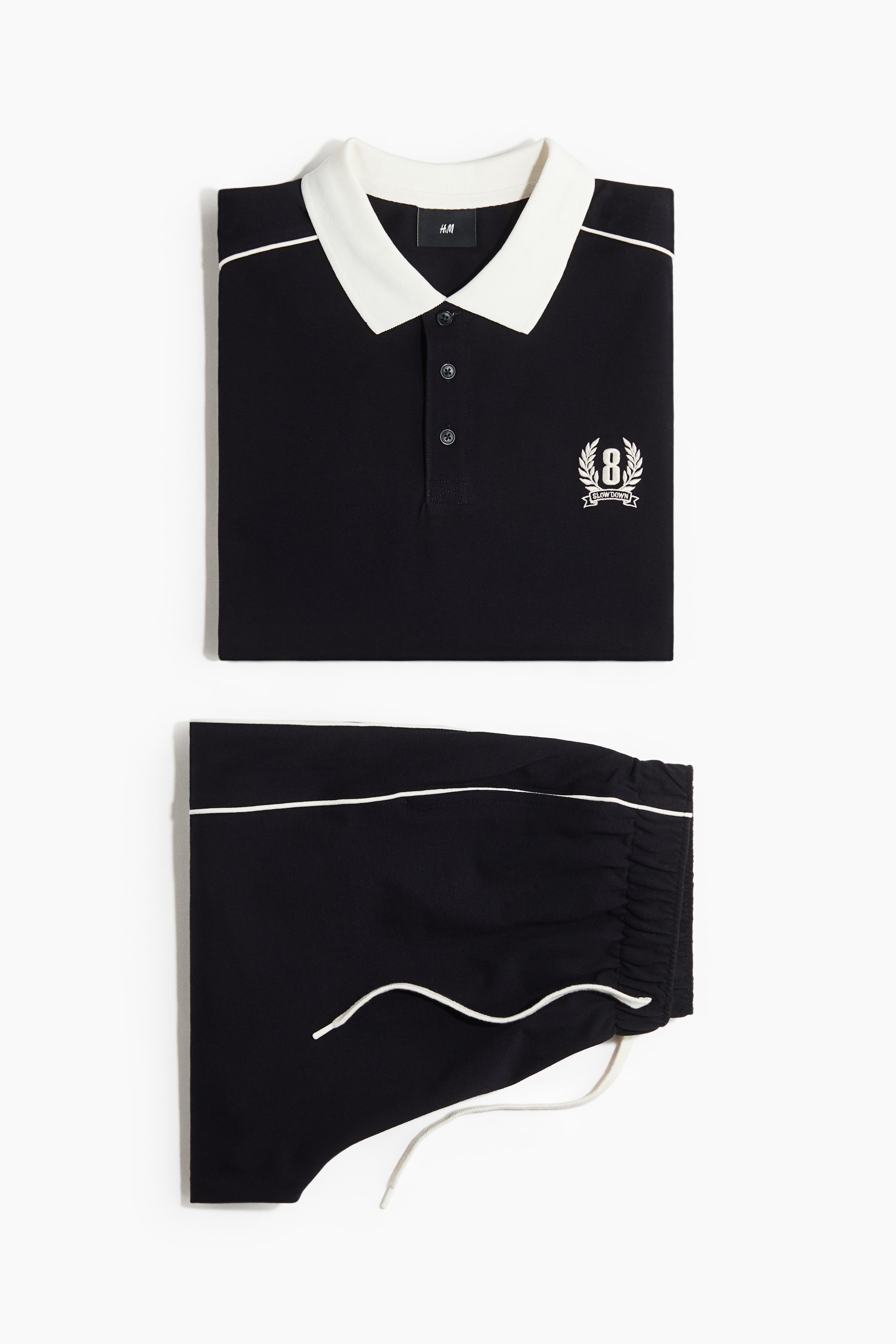 View larger image: A black polo shirt is displayed folded at the front, showcasing a white collar, a three-button placket, white piping along the shoulders and across the chest, and a white embroidered '8' emblem with 'SLOW DOWN' on the left chest. Coordinating black shorts are shown folded beneath the shirt, featuring white piping along the waistband, white drawstrings emerging from the front, and gathered leg openings.