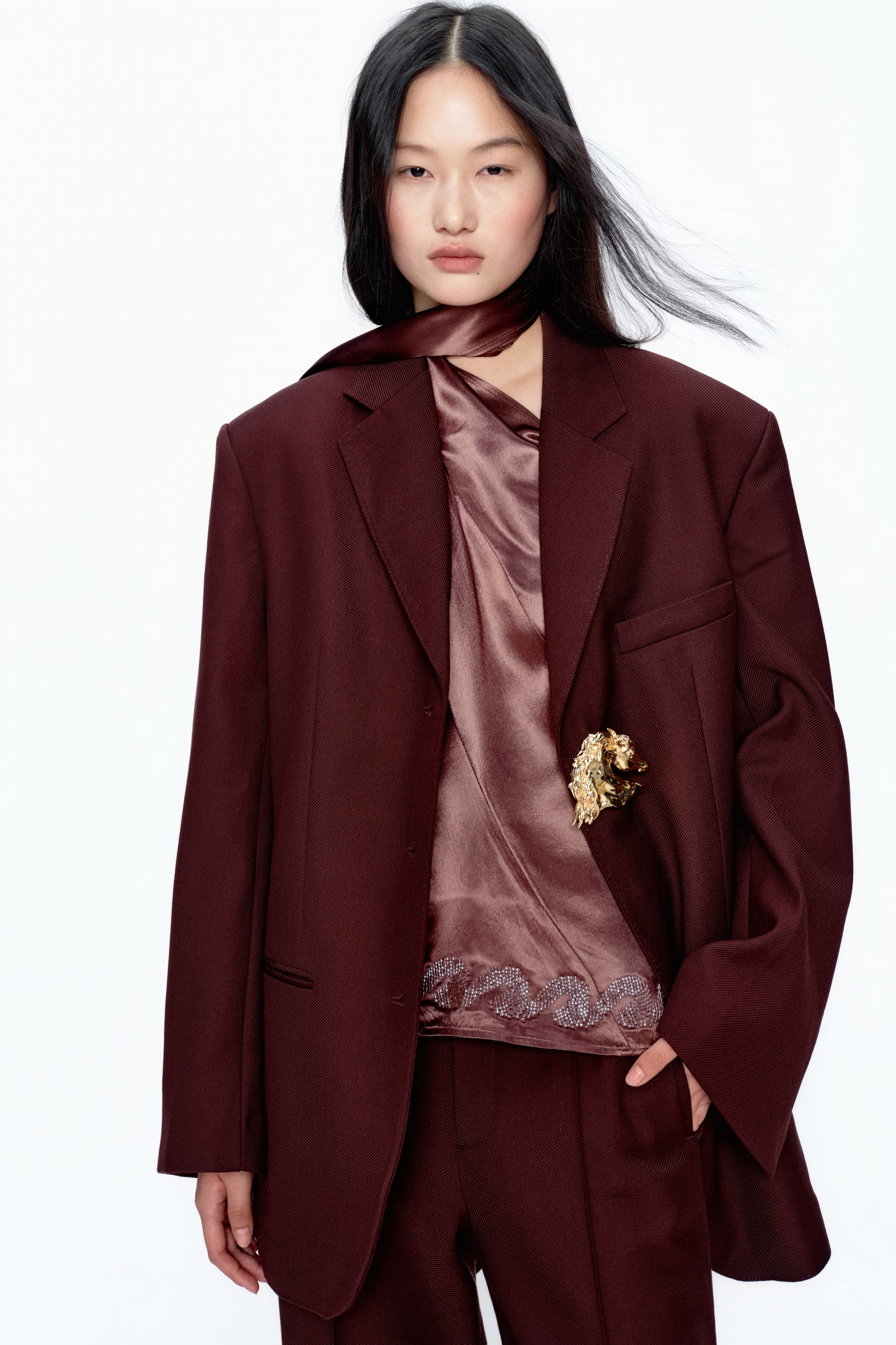 View larger image: A burgundy oversized blazer with structured shoulders and notch lapels is shown from a slight front-right angle, styled over a draped, satin-like top with an embellished wavy hem. A gold-tone horse head brooch accents the blazer’s left lapel, while matching burgundy trousers complete the look.