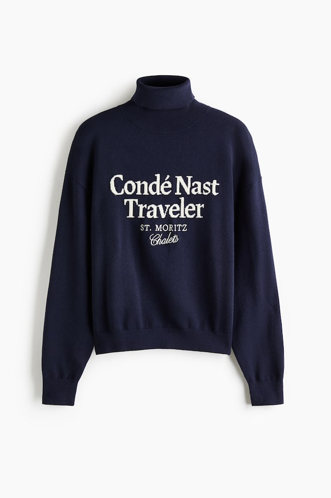 A navy blue turtleneck sweater is shown from the front, featuring white embroidered lettering on the chest that reads "Cond� Nast Traveler ST. MORITZ Chalets". The sweater is designed with a ribbed fold-over collar, dropped shoulders, and ribbed cuffs and hem. - 1