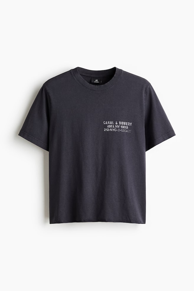A dark gray t-shirt is presented from the FRONT, featuring a ribbed crew neck and short sleeves. A faded light gray text graphic, reading "CANAL & BOWERY SOHO, NY 10013 212-NYC-GALLERY," is visible on the upper left chest. - 1