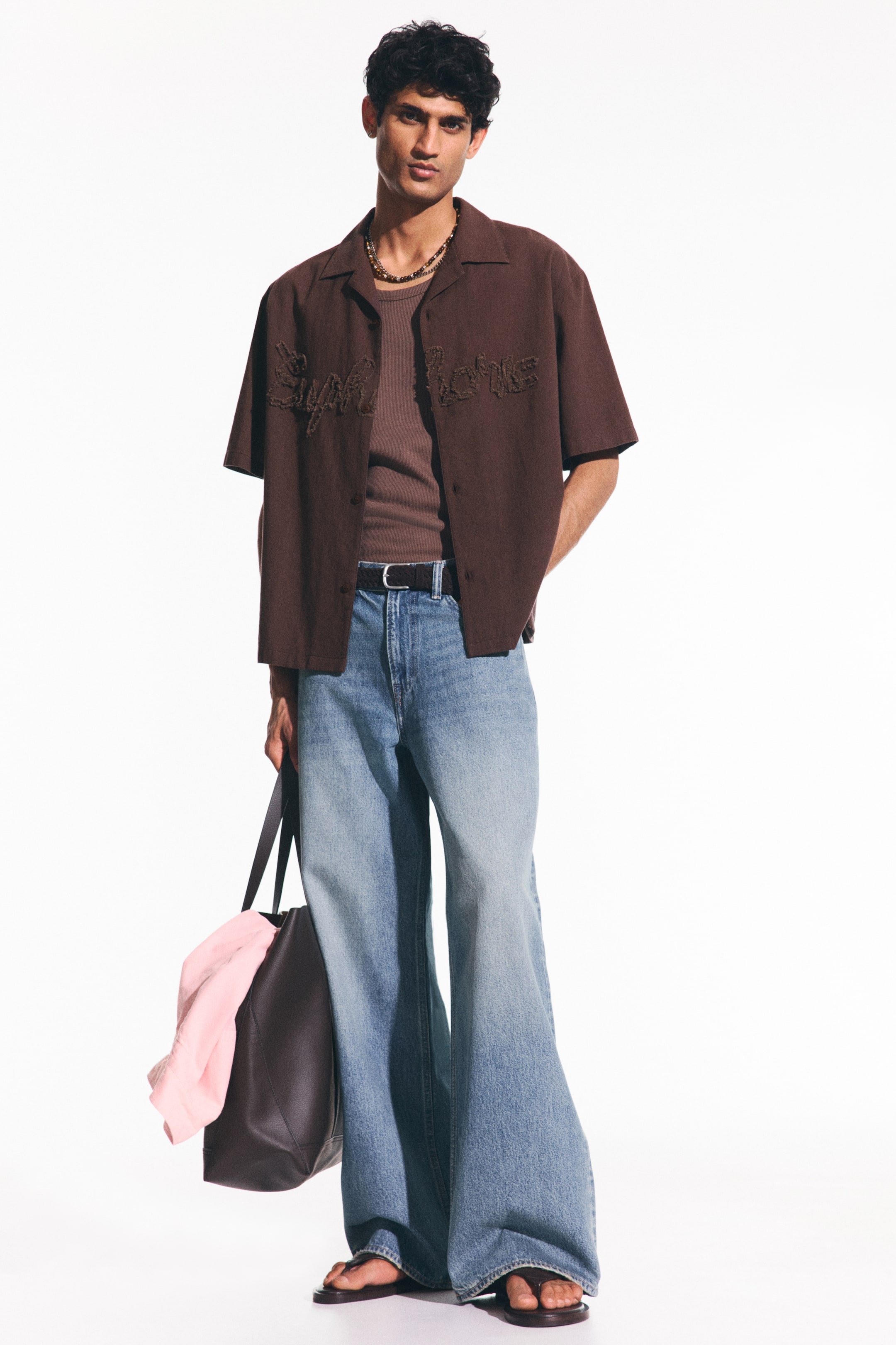 View larger image: The model is sporting a brown short-sleeved shirt featuring a resort collar and an embroidered motif on the front, styled open over a dark t-shirt. A beaded necklace can also be seen adorning the model's neck.