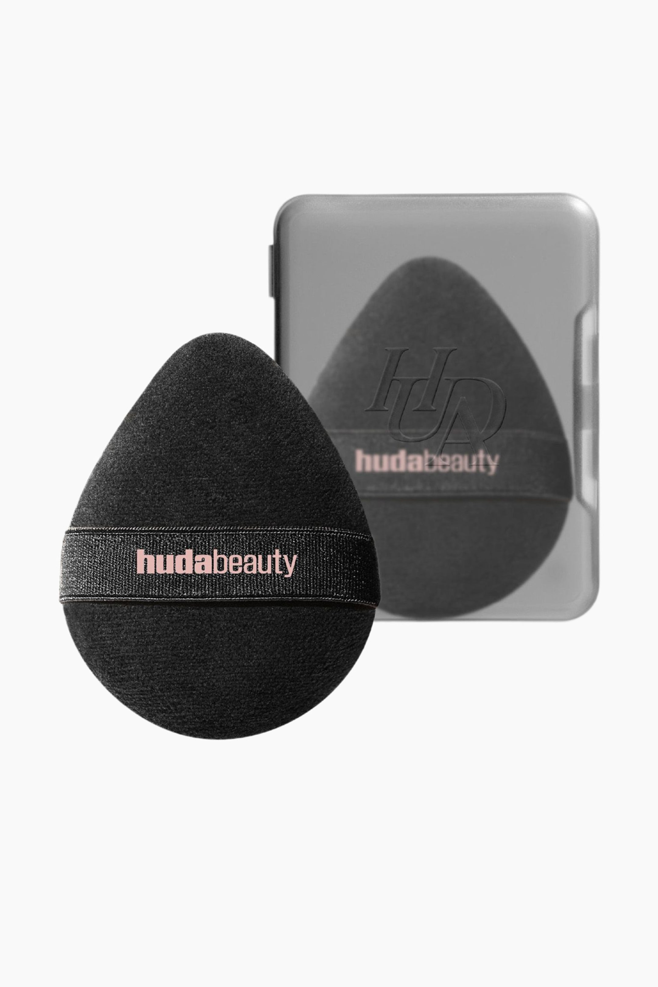 Näytä suurempi kuva: Musta, pisaranmuotoinen meikkipuffi on suunnattu eteenpäin, hieman oikealle kallistettuna, ja siinä on musta kuminauha, jossa on "hudabeauty"-teksti vaaleanpunaisena. Sen takana näkyy läpikuultava harmaa kotelo, jossa on toinen identtinen musta puffi, jossa myös näkyy merkin nimi.