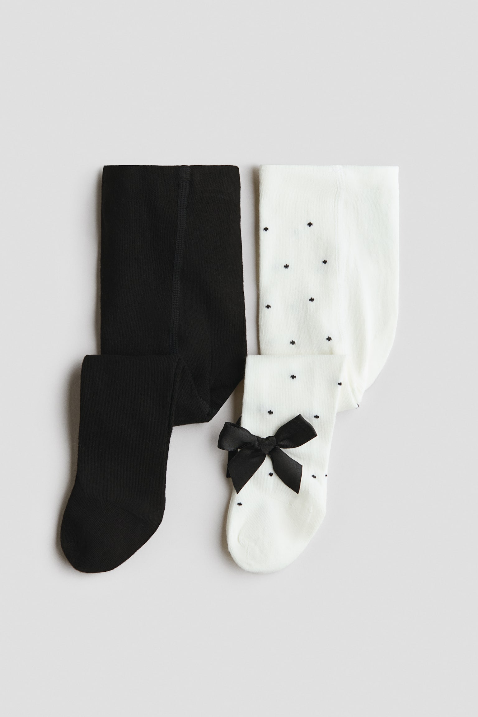 Two pairs of fine-knit cotton-blend tights are lying flat, with one pair in solid black and the other in white. The white pair is featuring a pattern of small black dots and a decorative black satin bow near the ankle.