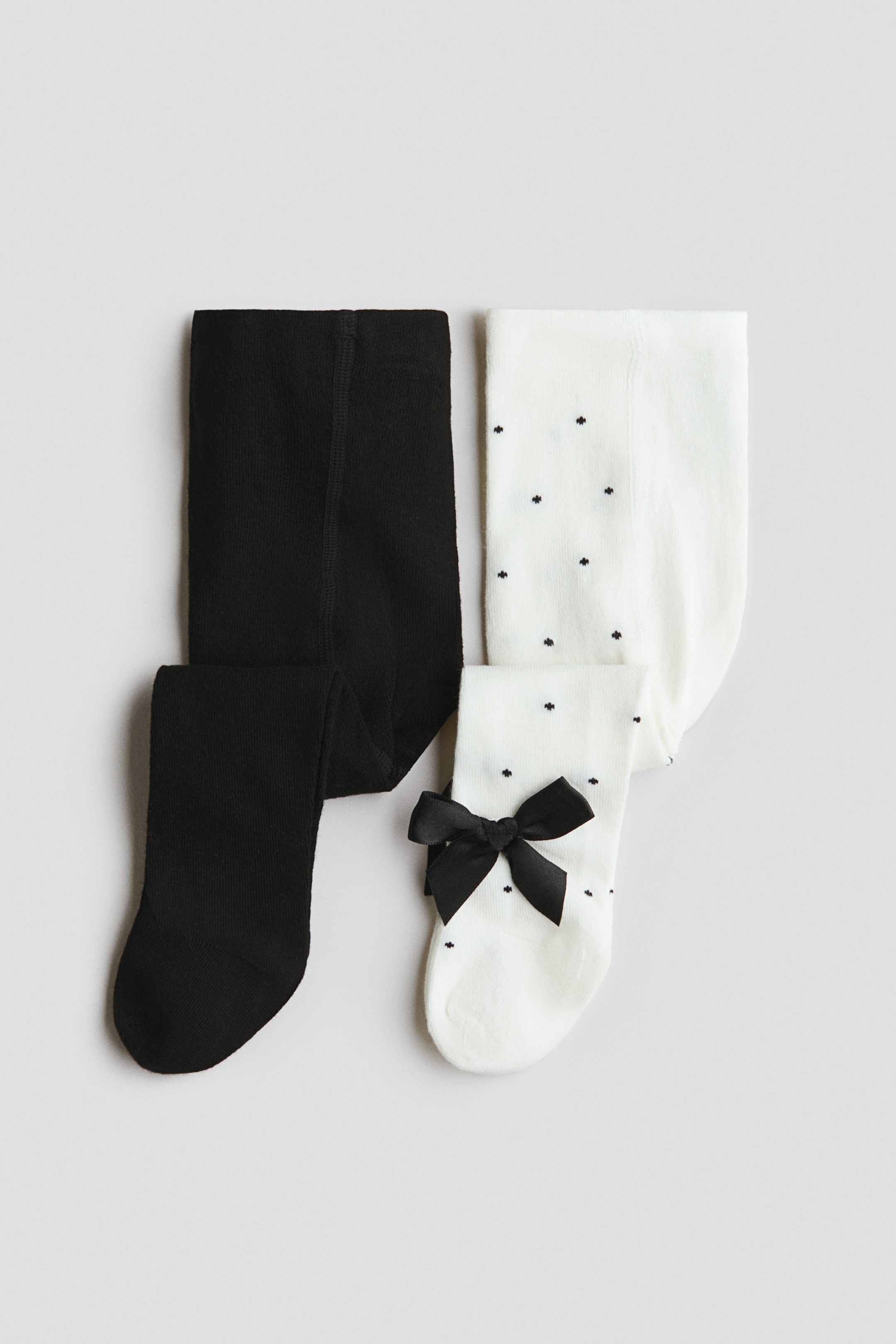 View larger image: Two pairs of fine-knit cotton-blend tights are laid flat, with one pair in solid black and the other in white. The white pair features a pattern of small black dots and a decorative black satin bow near the ankle.