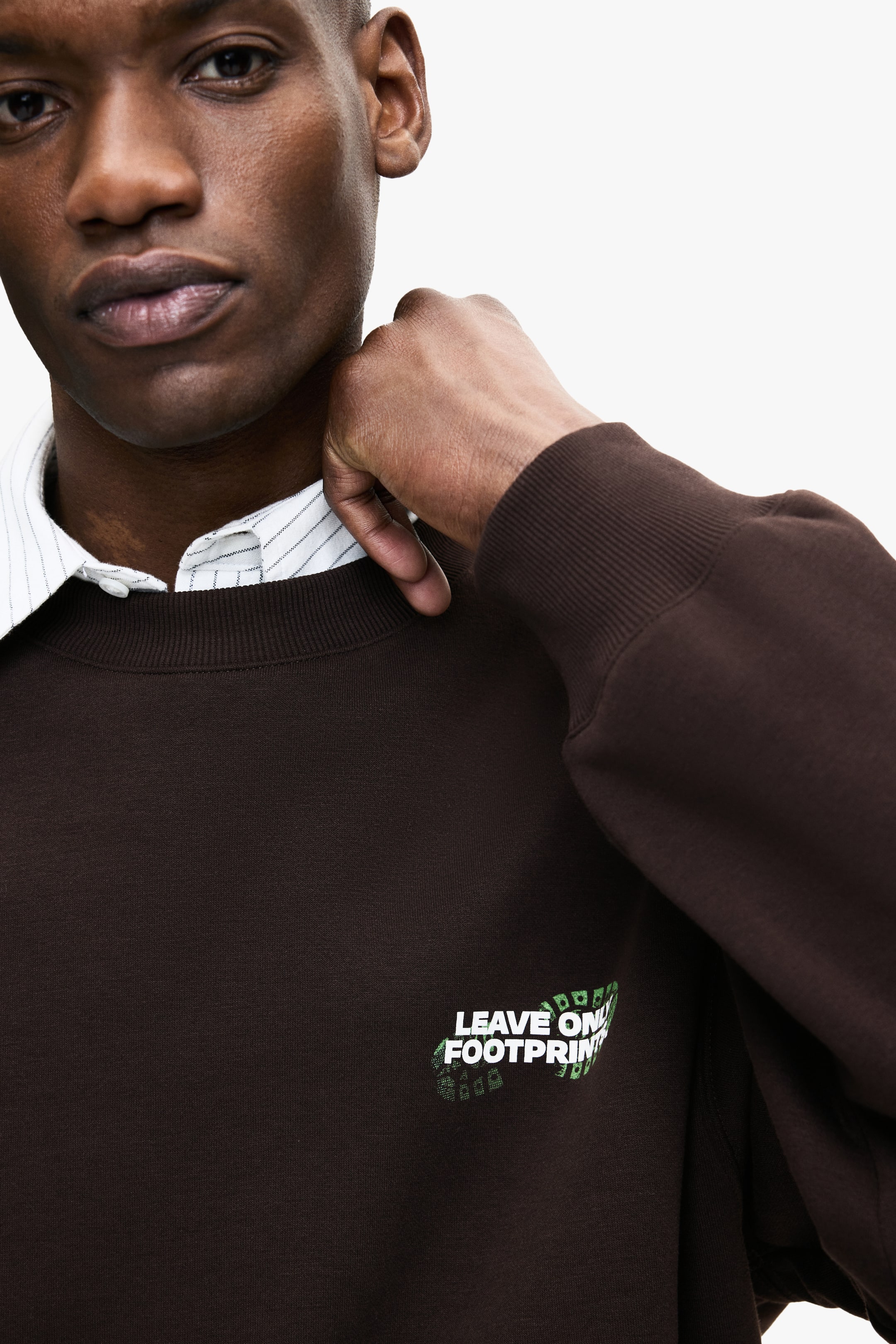 Grösseres Bild ansehen: Ein brauner Rundhals-Sweatshirt wird getragen, mit einem weißen und grünen "LEAVE ON FOOTPRINT"-Druck auf der Brust. Das Model passt den Halsausschnitt des Sweatshirts an, wobei ein weiß gestreiftes Hemd mit Kragen sichtbar ist.