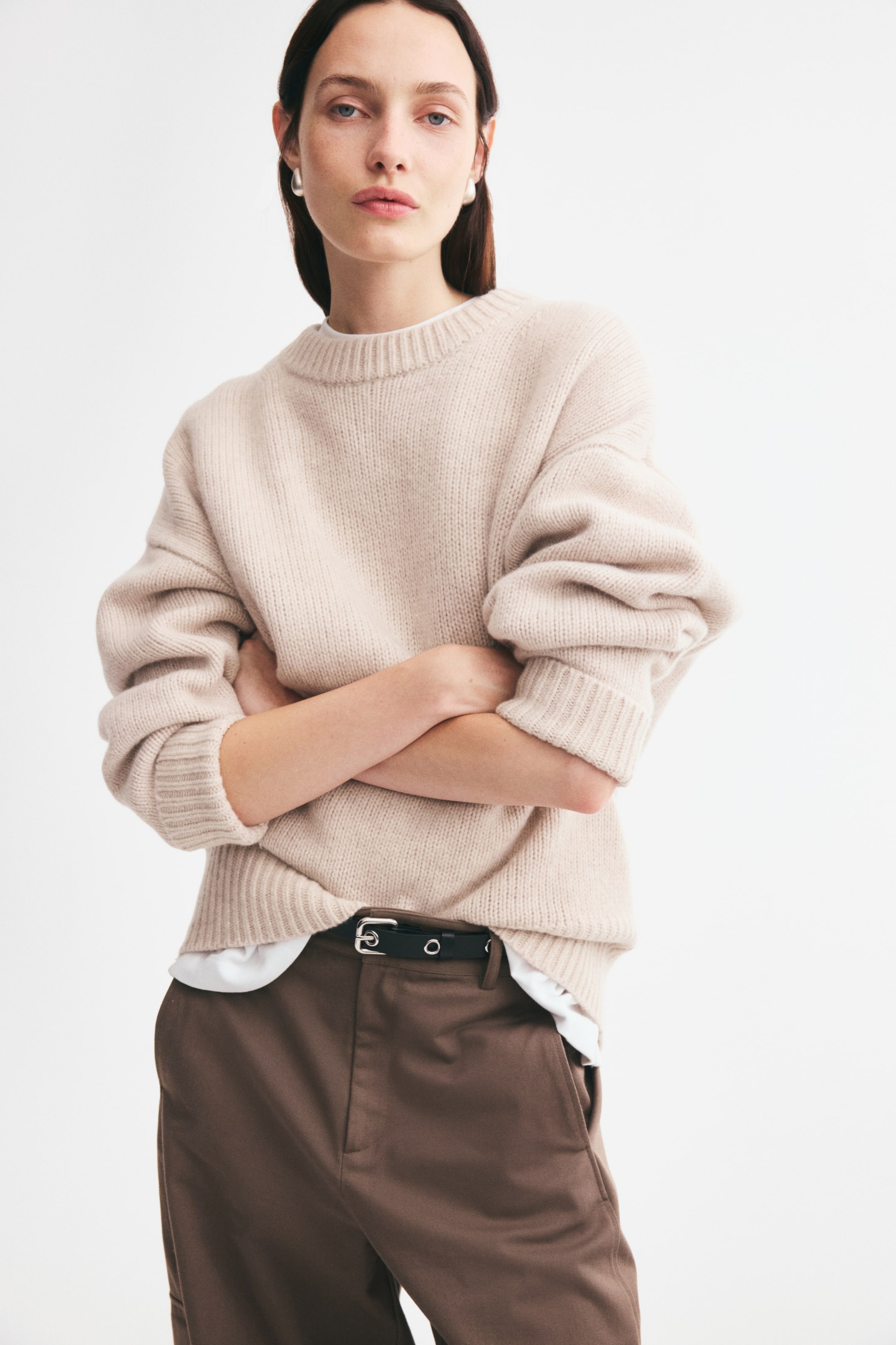 Agrandir l'image: Un pull en maille surdimensionné beige clair, doté d’un col rond côtelé, d’épaules tombantes et de poignets côtelés, est porté par le mannequin debout, les bras croisés. Le pull est superposé à un t-shirt blanc et associé à un pantalon marron.