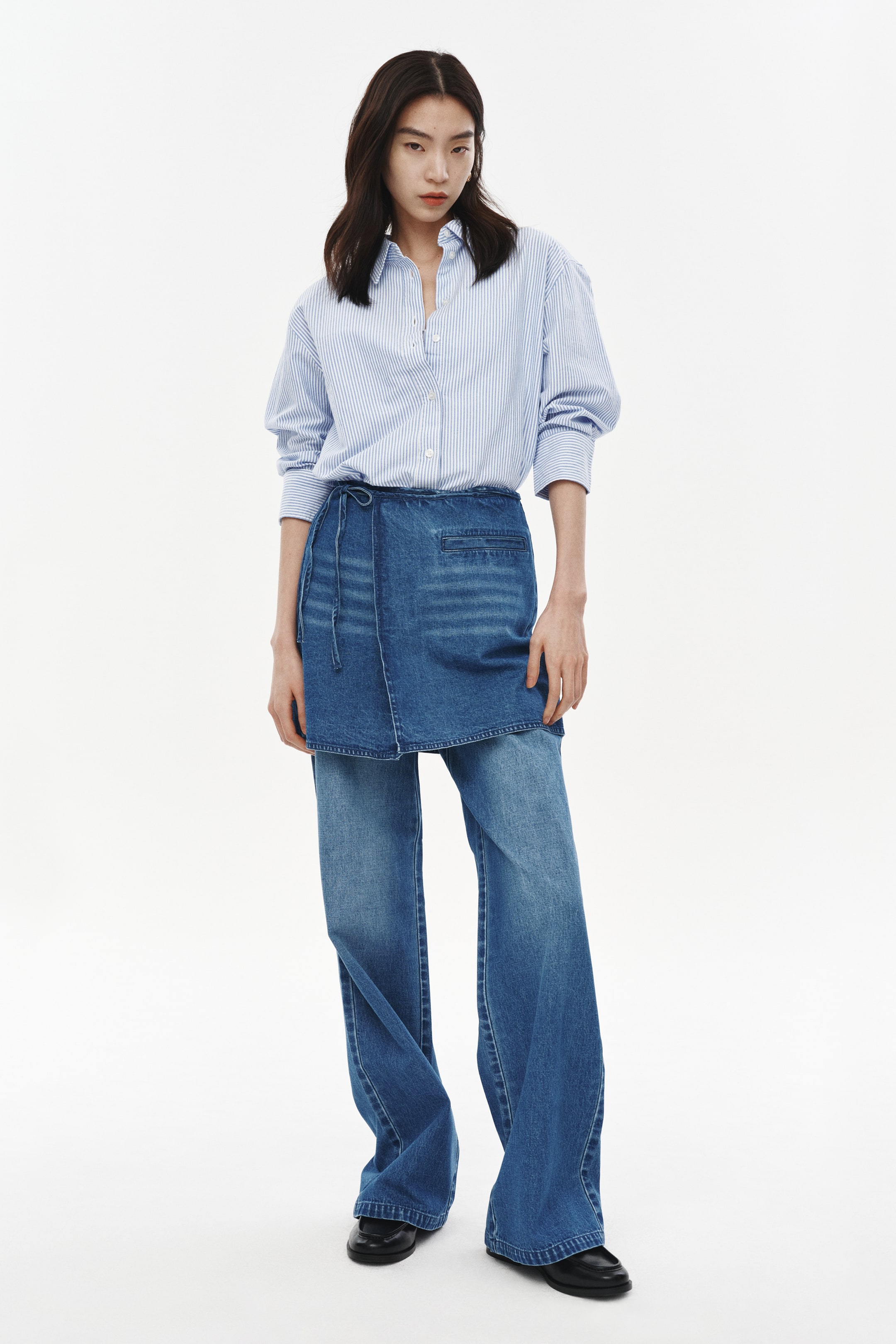 View larger image: A light blue and white striped button-down shirt is pairing with wide-leg blue denim jeans and a blue denim wrap skirt. The shirt is featuring a collar and buttoned cuffs. The denim wrap skirt is tying at the waist and layering over the jeans, which are presenting a relaxed, wide-leg fit.