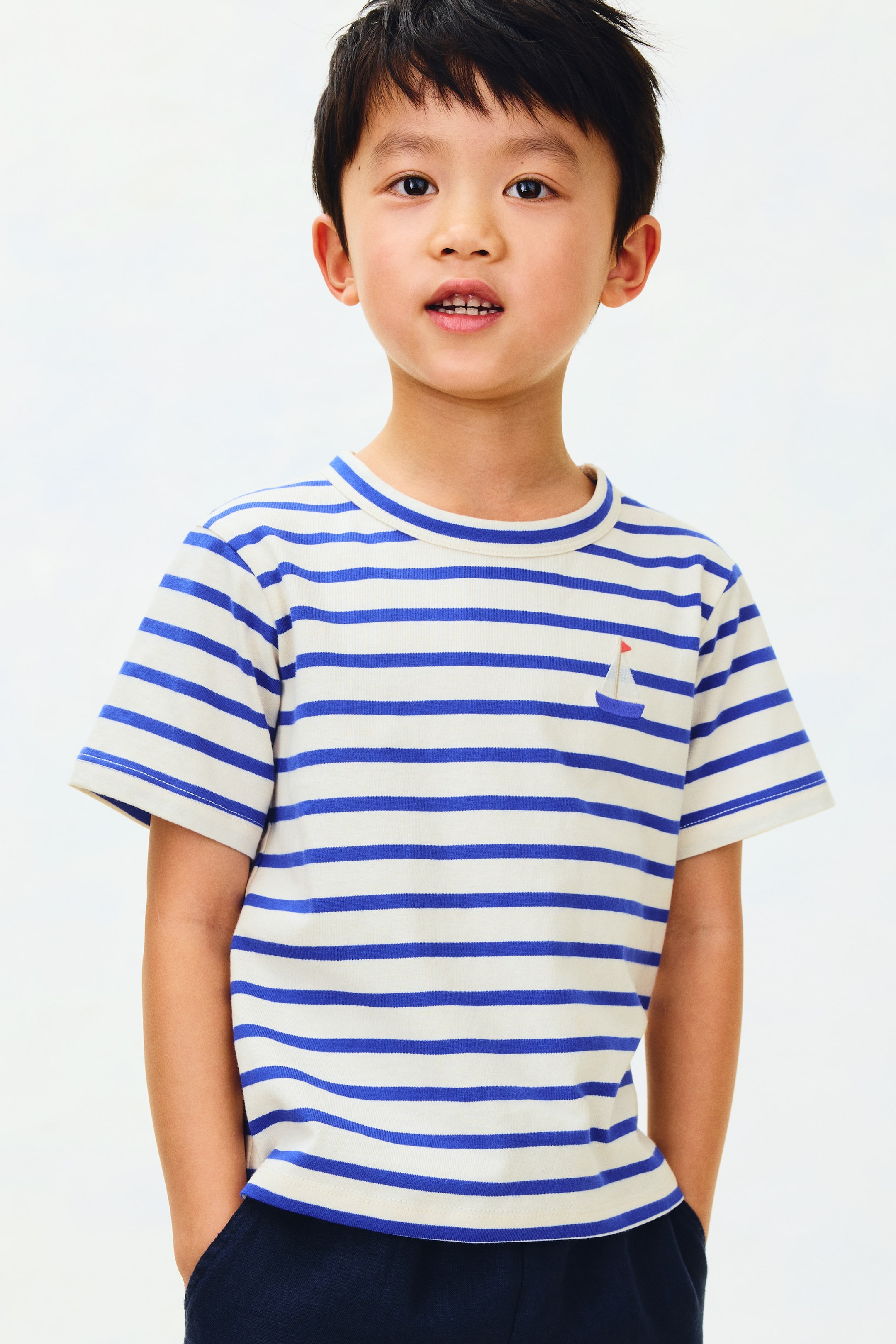 Se større billede: En kortærmet t-shirt med vandrette off-white og blå striber, en rund halsudskæring og en lille blå sejlbåd broderet på venstre bryst.