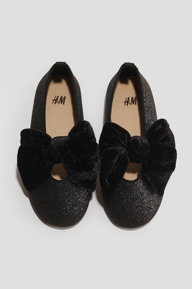 A pair of black glittery ballet flats is showcasing a large decorative black velvet bow secured across the front of each shoe. The shoes feature a rounded toe silhouette, black elasticised trim, and a beige insole. - 1