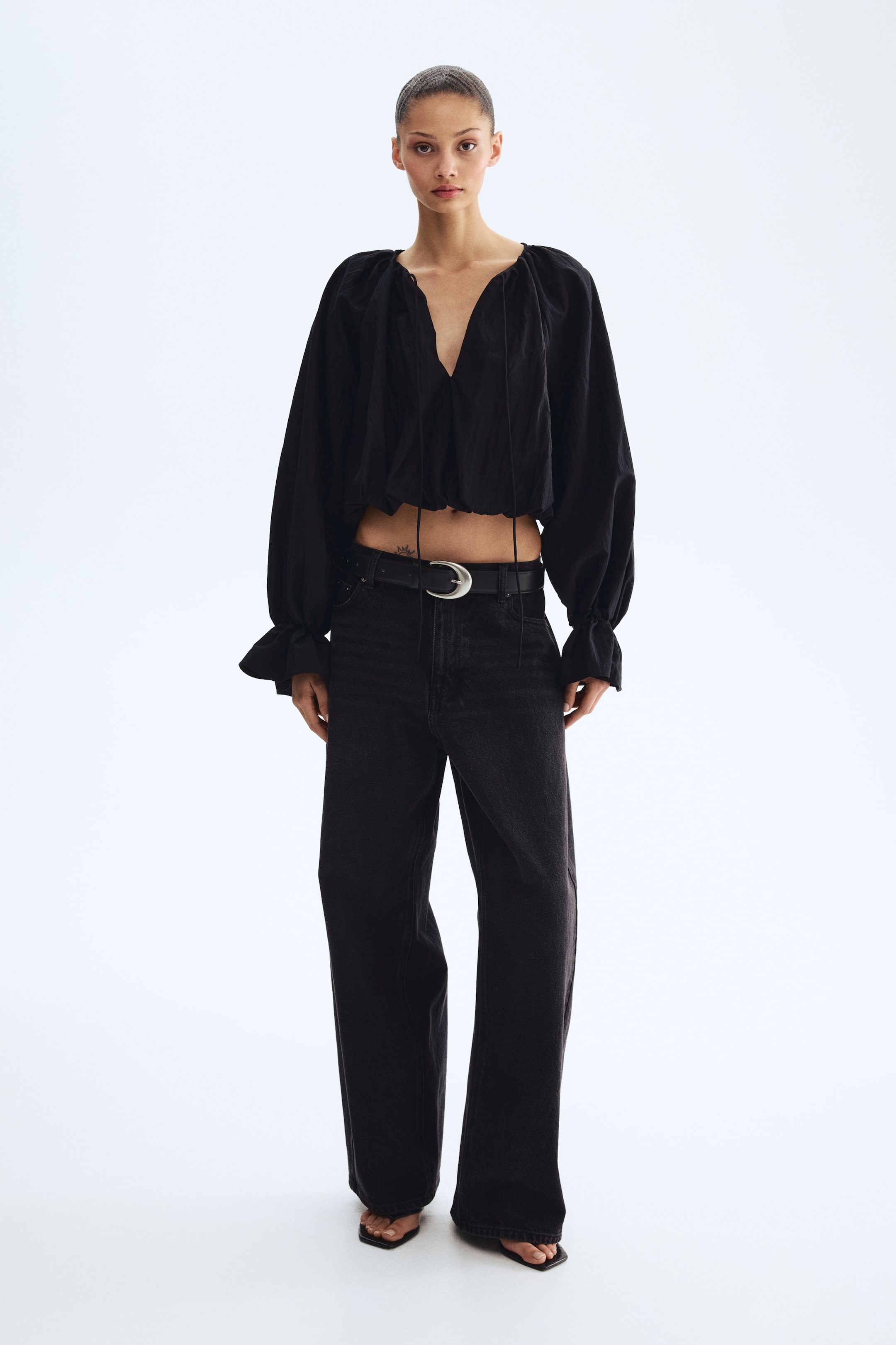 Voir une image plus grande: Un haut blouson court froissé noir, doté d'un col en V avec cordons de serrage et de manches volumineuses avec poignets à volants, est porté. Il est associé à un jean noir délavé à jambes larges, cintré par une ceinture noire avec une boucle ovale argentée et stylisé avec des sandales à bout carré noir.