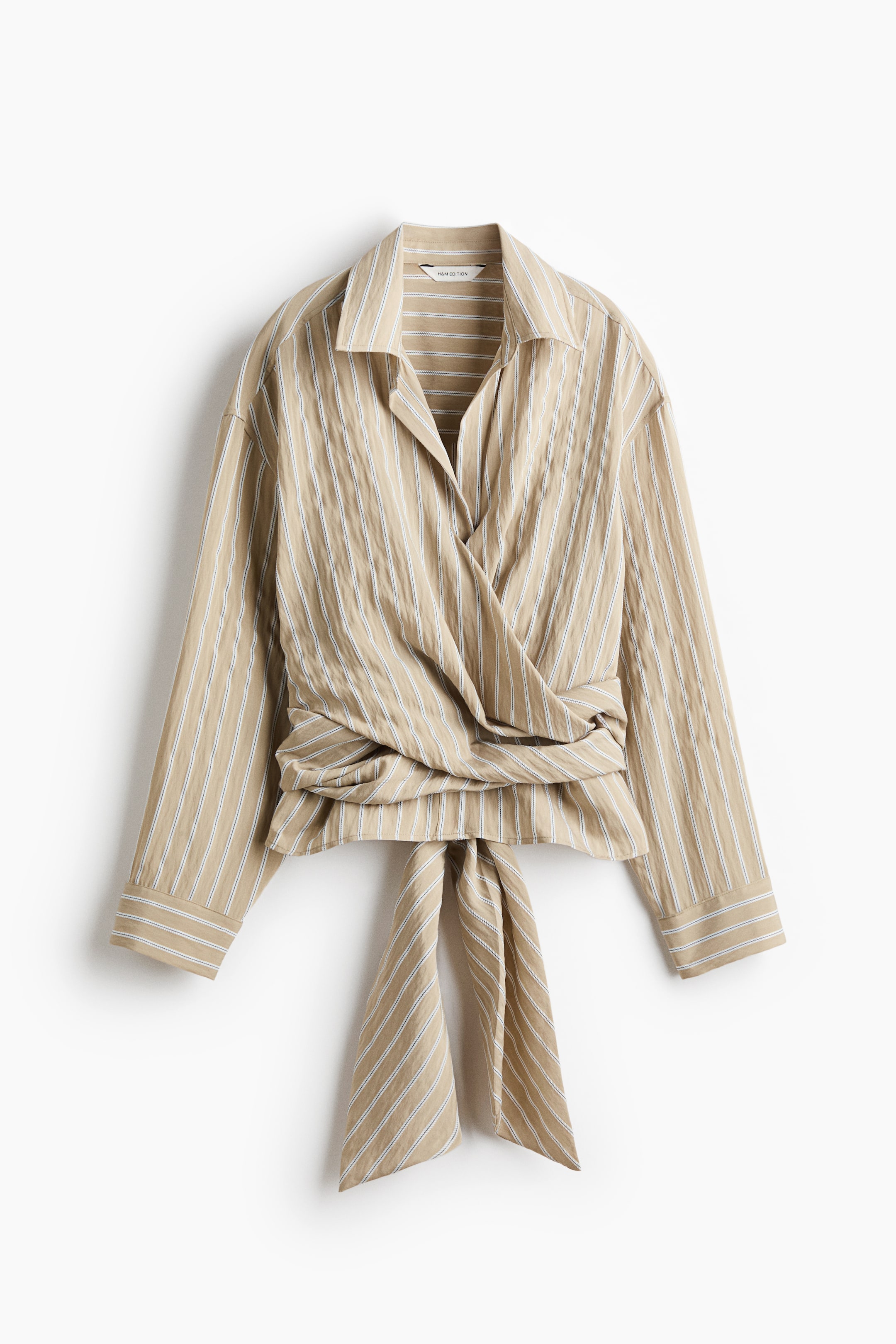 Grösseres Bild ansehen: Eine beige gewebte Bluse mit weißen vertikalen Streifen wird in der Vorderansicht präsentiert. Sie verfügt über einen Kragen, überschnittene Schultern, lange Ärmel mit geknöpften Manschetten und breite Wickelpartien, die sich vorne kreuzen, die Taille betonen und am unteren Vorderteil zu einem Knoten gebunden werden.