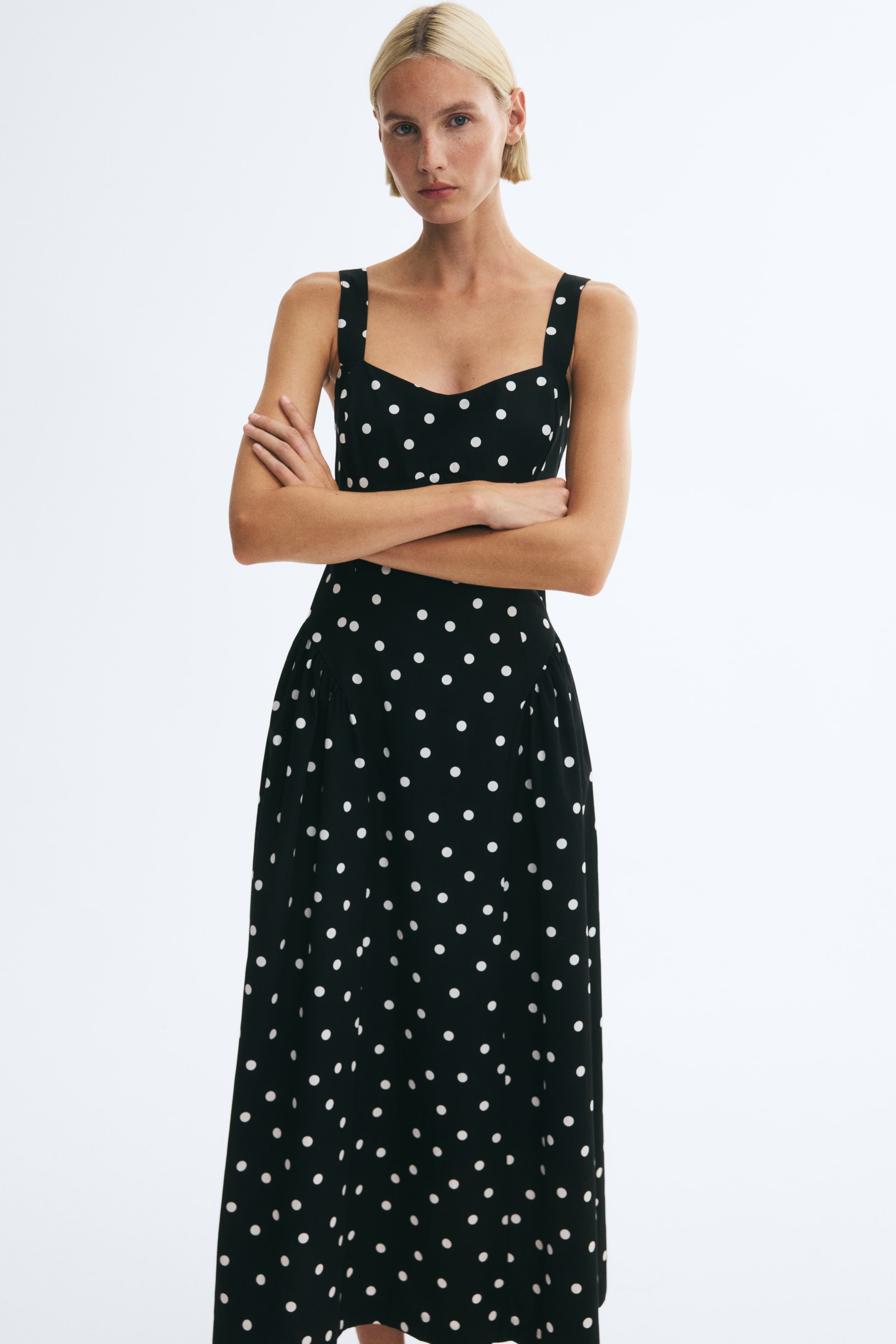 Größeres Bild ansehen: Ein schwarzes, knöchellanges Kleid mit weißem Polka-Dot-Muster wird von dem Model getragen, das mit verschränkten Armen steht. Das Kleid verfügt über einen Herzausschnitt mit breiten Trägern, ein tailliertes Oberteil und einen ausgestellten Rock mit gerafften Nähten über den Hüften.