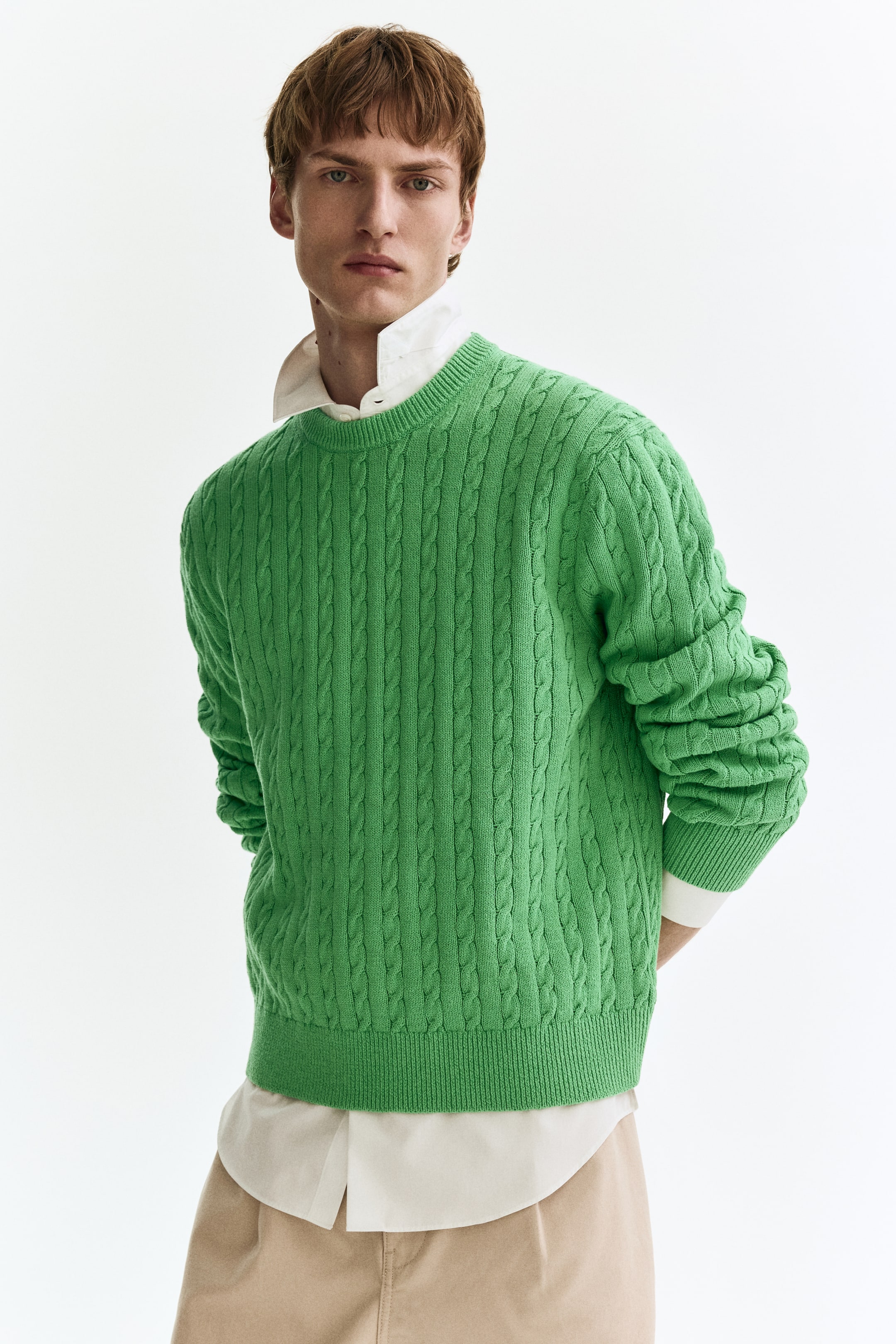 Visualizza immagine più grande: Un maglione a trecce di un vivace verde brillante presenta un girocollo, polsini a coste e un orlo a coste. È indossato sopra una camicia bianca con colletto, con il colletto e l'orlo arrotondato della camicia visibili sotto, ed è presentato da un'angolazione frontale sinistra.