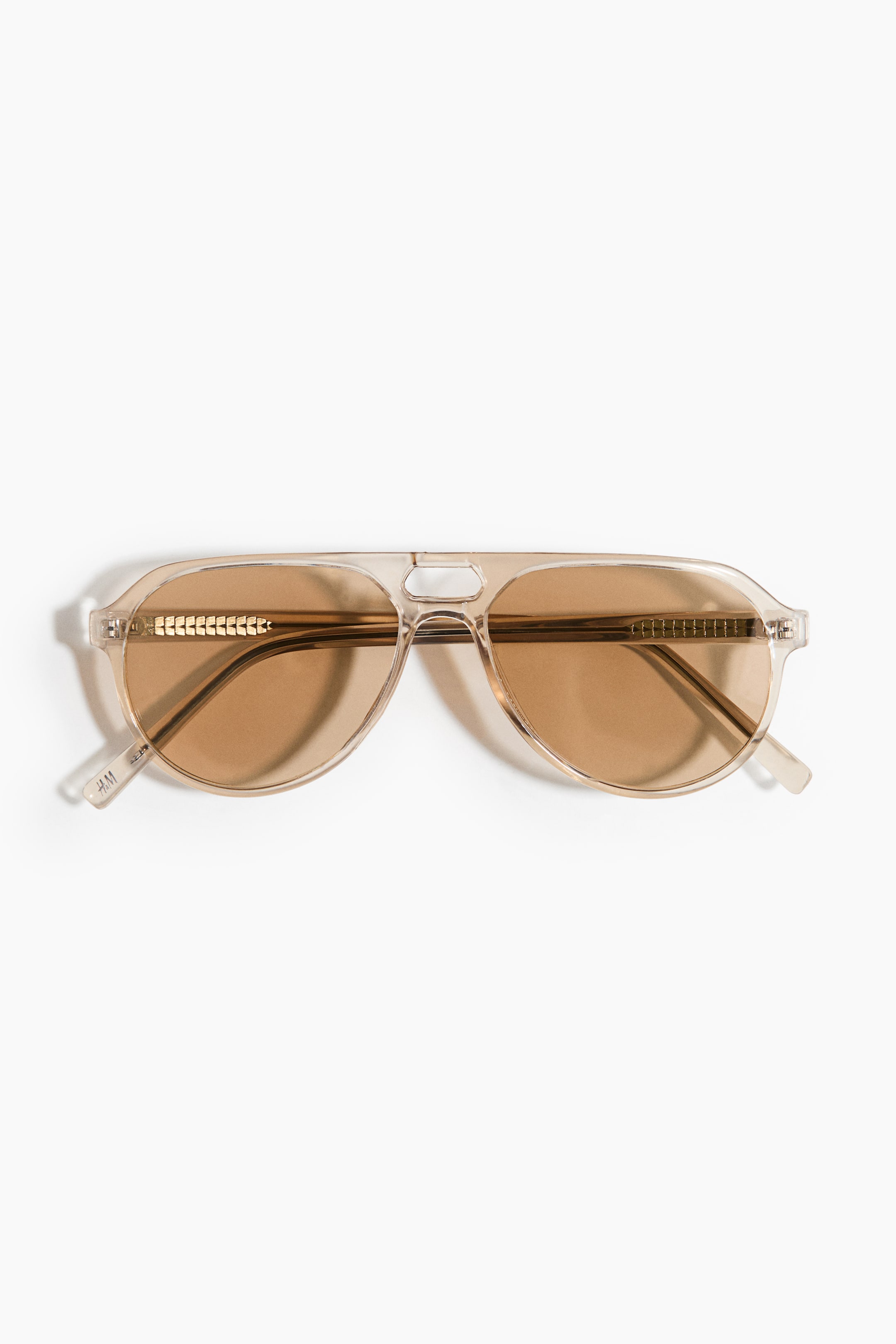 Größeres Bild anzeigen: Die Sonnenbrille ist frontal ausgerichtet und verfügt über einen transparenten, hellbraunen Rahmen im Aviator-Stil mit braun getönten Gläsern. Der Rahmen zeigt einen Doppelsteg, wobei der obere Steg eine sechseckige Aussparung aufweist. Goldfarbene, segmentierte Metallakzente sind an der Innenseite des Rahmens nahe den Scharnieren zu sehen, und auf dem linken Bügel ist ein „H&M“-Logo sichtbar.