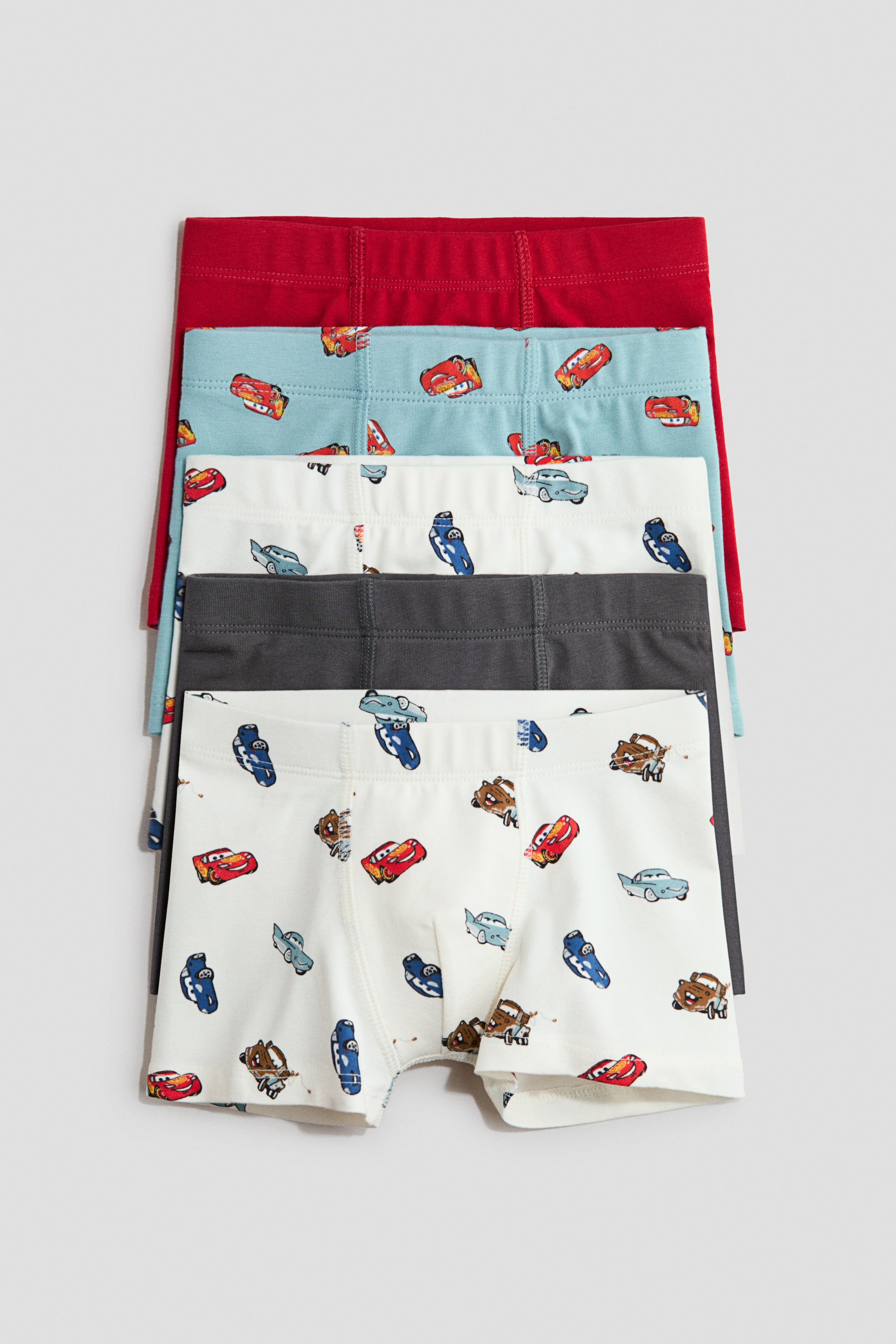View larger image: Five pairs of boys' boxer briefs are neatly arranged in a flat stack, showcasing an array of vibrant colors and playful patterns. From top to bottom, the stack reveals a solid red pair, followed by a light blue pair adorned with a repeating Disney Cars character motif, a white pair featuring a similar Cars design, a solid dark grey pair, and another white pair with the Cars print.