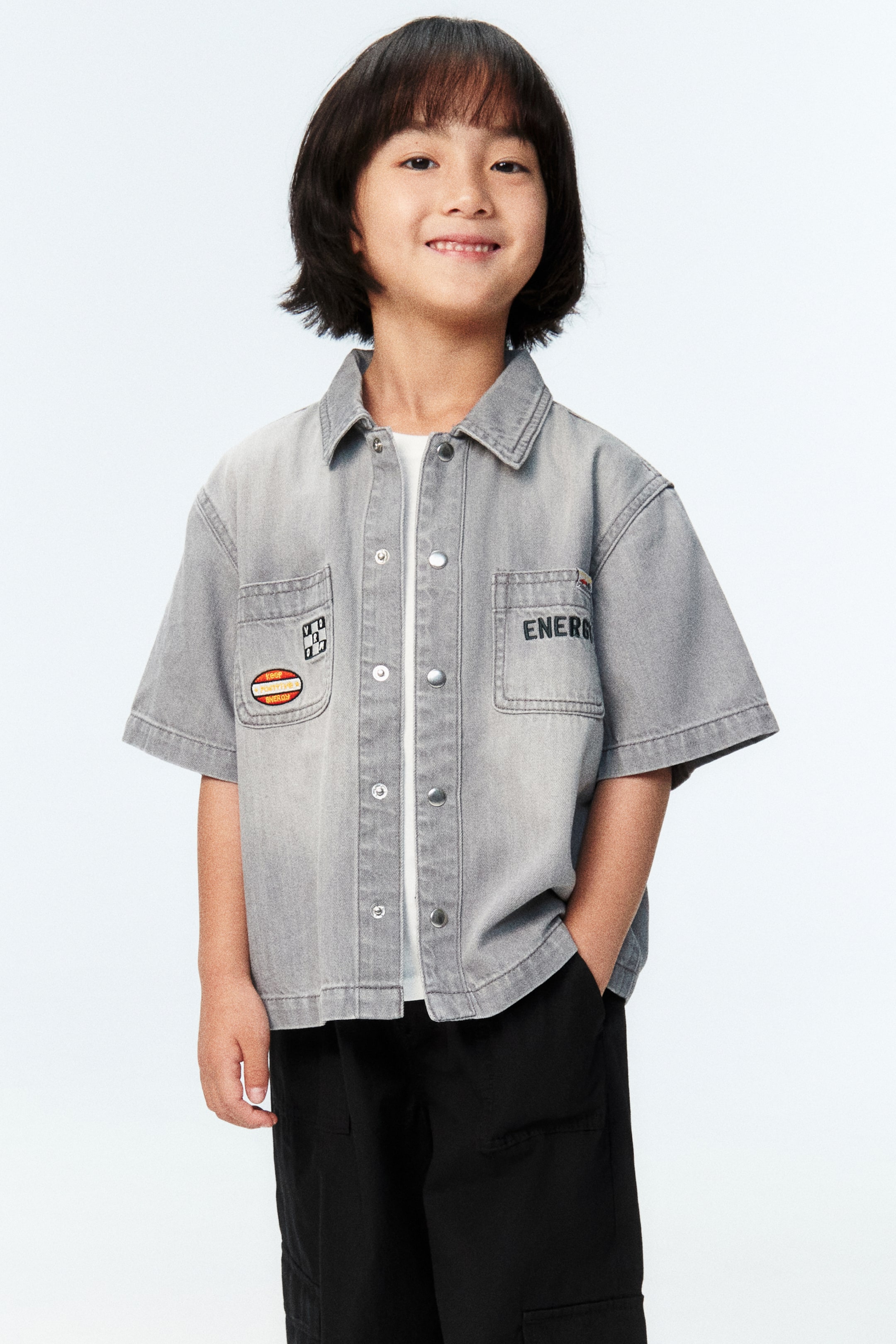 Xem ảnh lớn hơn: Người mẫu đang mặc một chiếc áo sơ mi denim màu xám ngắn tay, được phối cùng áo thun trắng bên trong. Chiếc áo sơ mi có cổ áo, nút bấm, hai túi ngực có nắp, các miếng trang trí, và thêu chữ "ENERGY".
