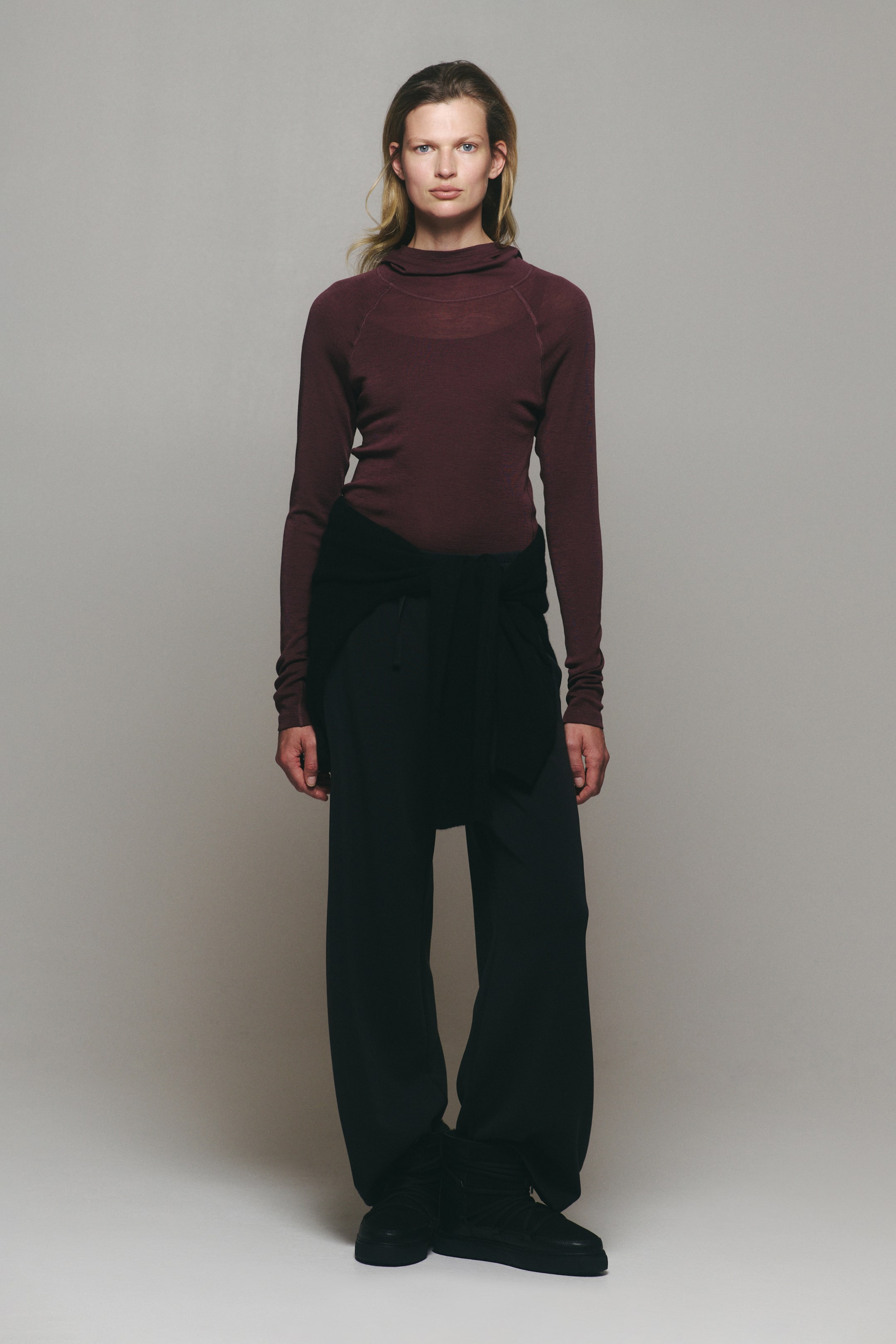 Agrandir l'image: Un haut de sport ajusté couleur bordeaux en jersey de laine, doté d’une capuche drapée, de manches raglan longues et de coutures flatlock, est porté par le mannequin. Le haut est assorti à un pantalon noir à jambes larges et à un pull noir noué à la taille.