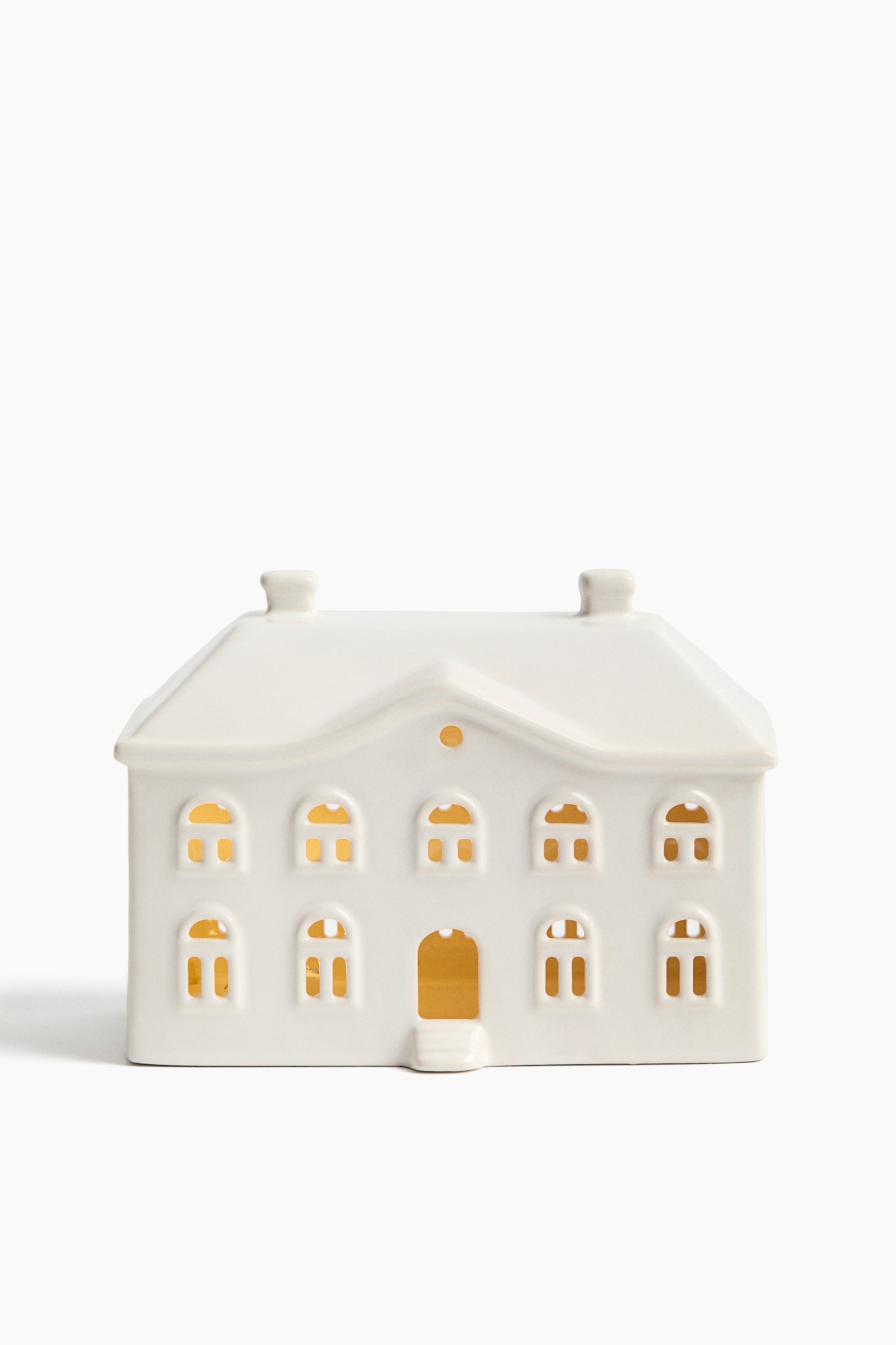 Se større billede: En hvid keramisk lanterne formet som et hus vender frontalt og har et skråt tag med to skorstenslignende strukturer. Facaden har flere udskårne vinduer i bue- og rektangulær form samt en central buet døråbning med trin, alt sammen med varmt lys, der skinner indefra.