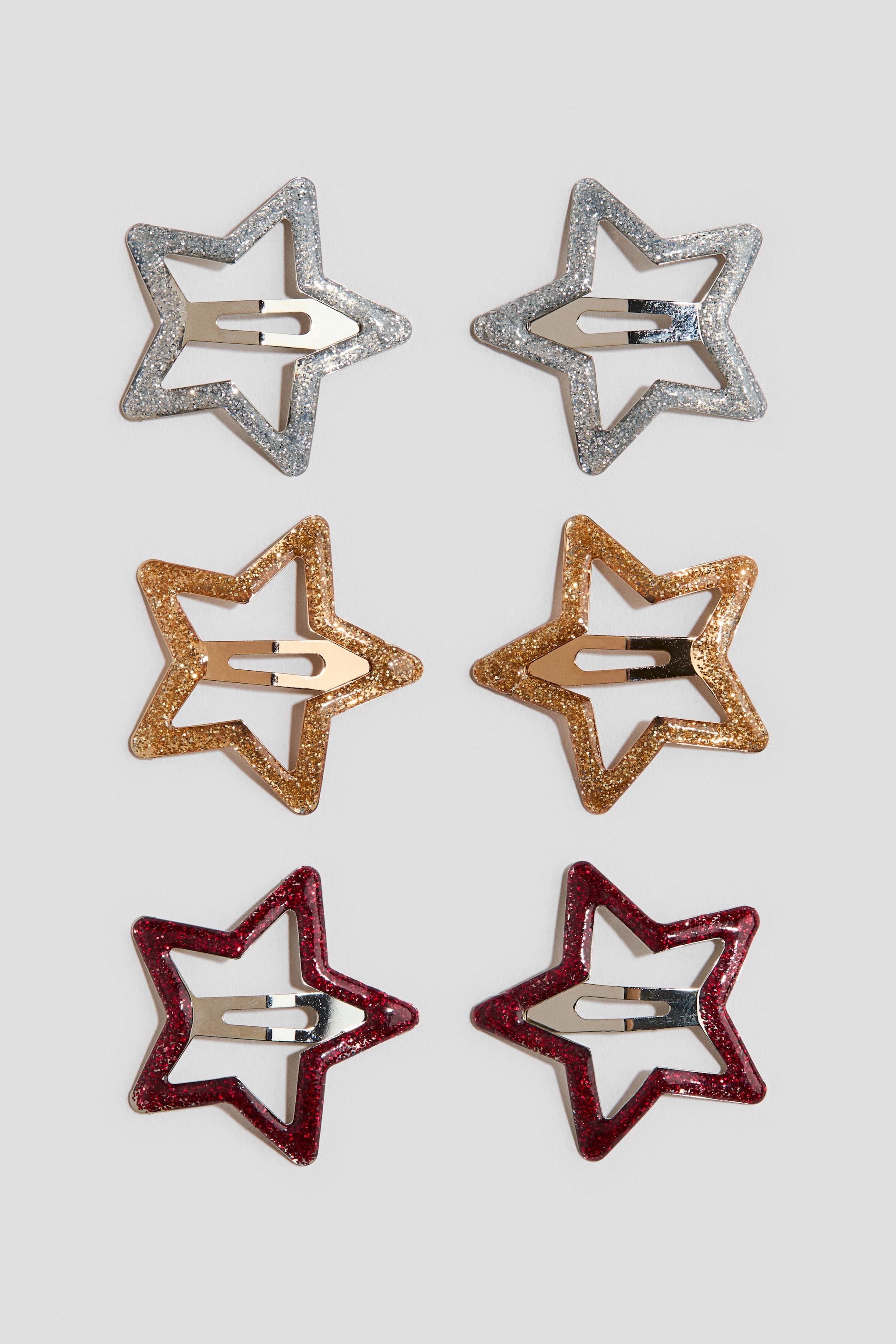 View larger image: Six star-shaped metal snap hair clips are shown from the front, arranged in pairs with silver, gold, and red glitter finishes. Each clip features an open-frame star design with a central metallic snap mechanism.