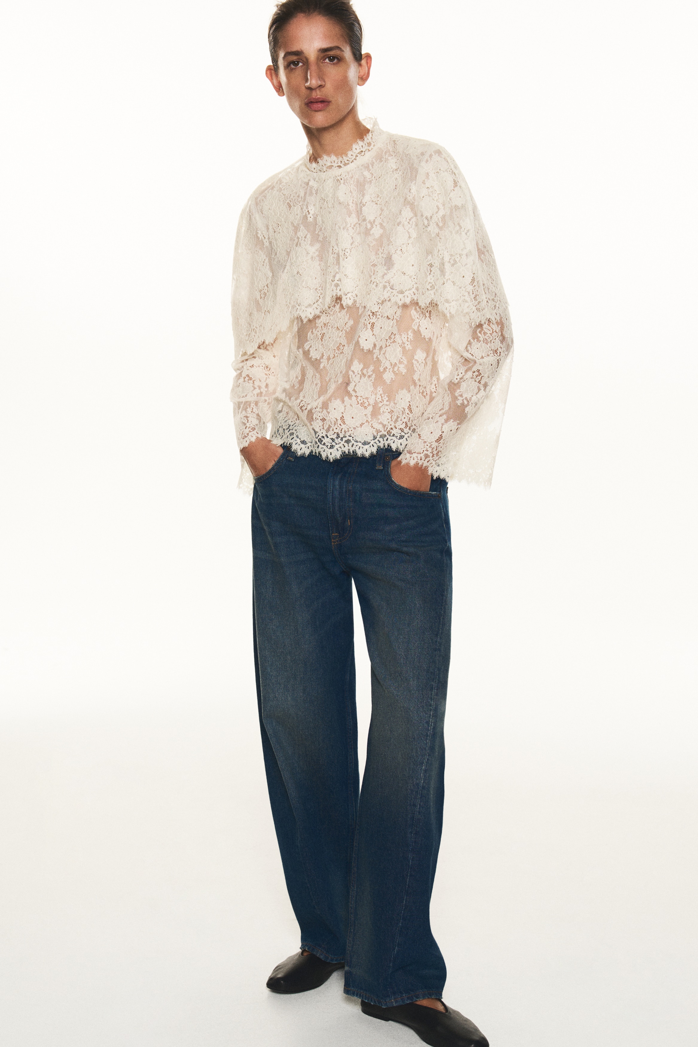 A front view showing a cream-coloured cropped lace blouse featuring a high stand collar, long wide sleeves, and a draped, cape-like overlay at the top. The entire piece is constructed from delicate floral lace and features scalloped, eyelash-stitch edges along the collar, cuffs, hem, and overlay.
