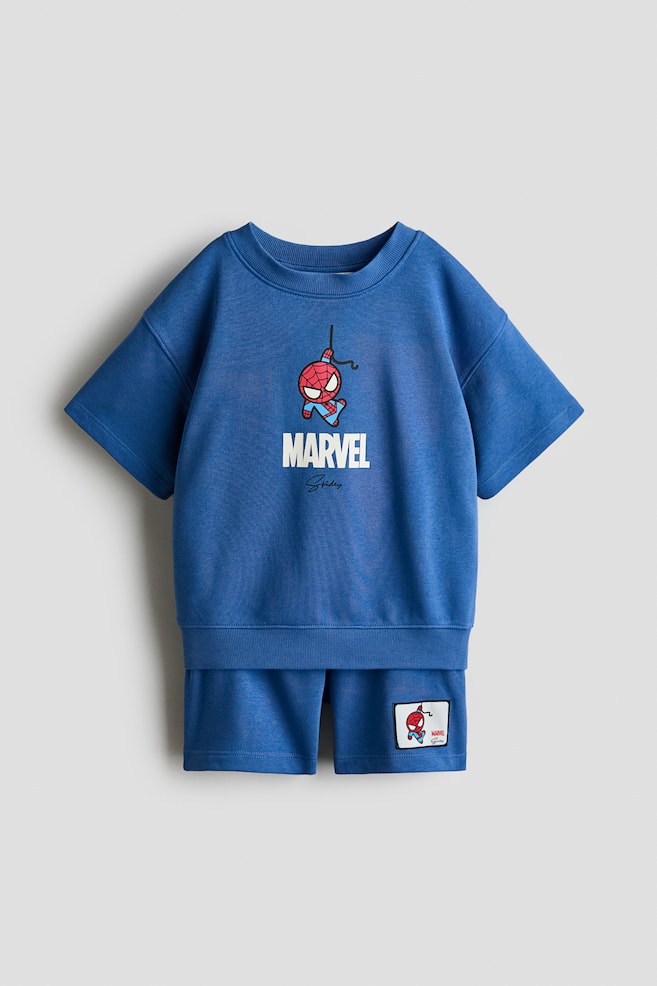 A blue set, shown from the front, features a T-shirt and shorts made from sweatshirt fabric. The T-shirt displays a relaxed fit with low, dropped shoulders, wide, short sleeves, and ribbing around the crew neckline and hem, together with a Spider-Man graphic and "MARVEL" text. The shorts feature an elasticated waist and a small Spider-Man patch on the right leg. - 1