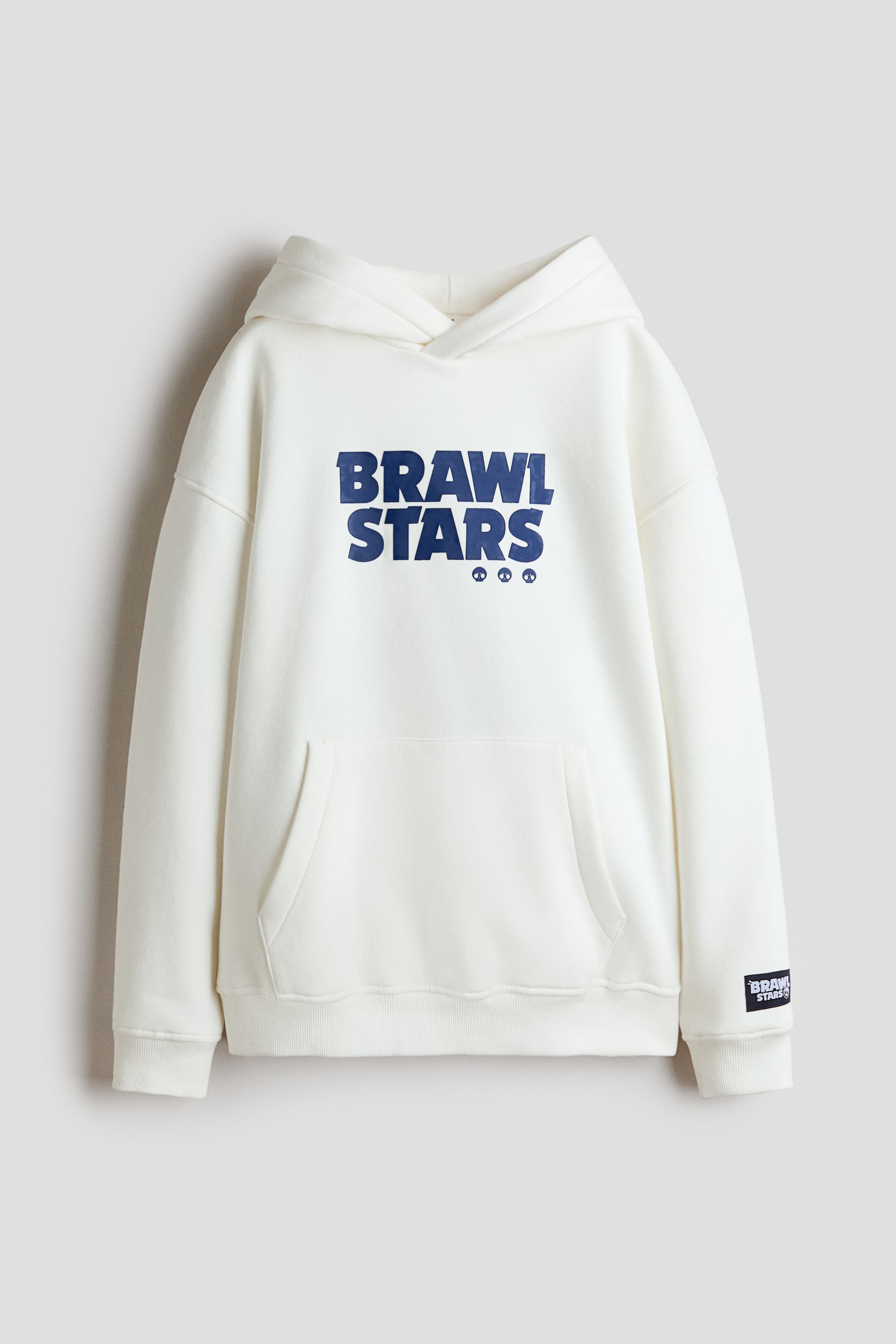 Visa större bild: En löst sittande, off-white huvtröja visas framifrån, med ett blått 'BRAWL STARS'-tryck och tre små dödskalleikoner nedanför. Den är designad med en fodrad huva, lågt nedfallna axlar, en känguruficka, långa ärmar med ribbade muddar, en ribbad nederkant och en svart 'BRAWL STARS'-patch på vänster ärm.