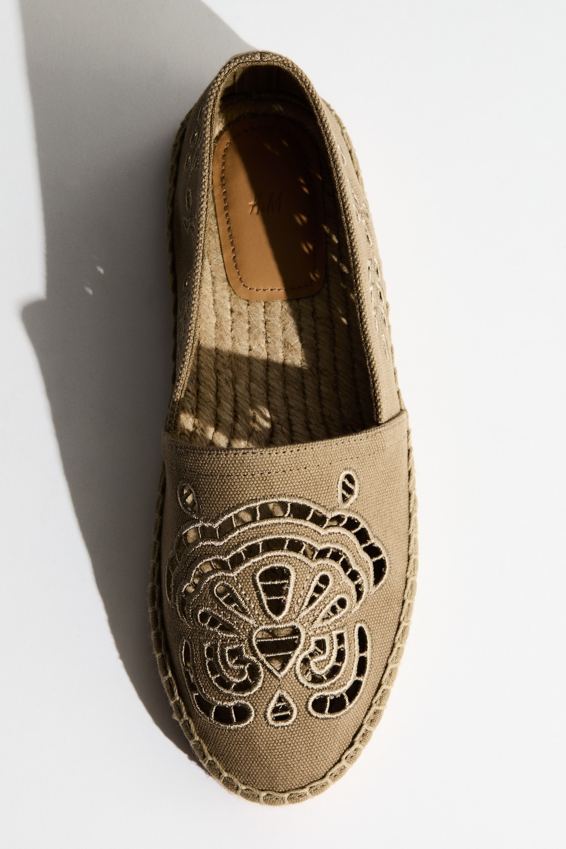 View larger image: An espadrille shoe features a khaki canvas upper adorned with an intricate cut-out embroidery design on the vamp, depicting a stylized animal face. It is detailed with a braided jute sole and perforated patterns along the sides, while a brown leather patch is visible inside the heel.