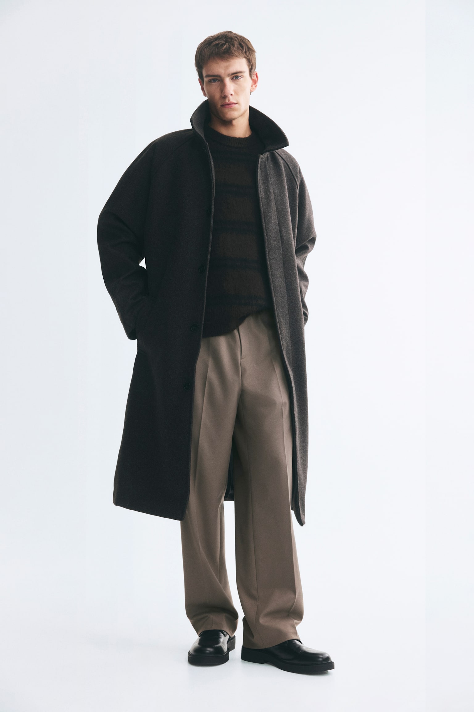 Tailored joggers in a taupe woven fabric are being worn by the model, featuring a relaxed silhouette and sharp creases running down the front of the legs. The trousers are styled with a dark, horizontally striped knit sweater, a long dark-coloured overcoat, and black leather shoes. - 1