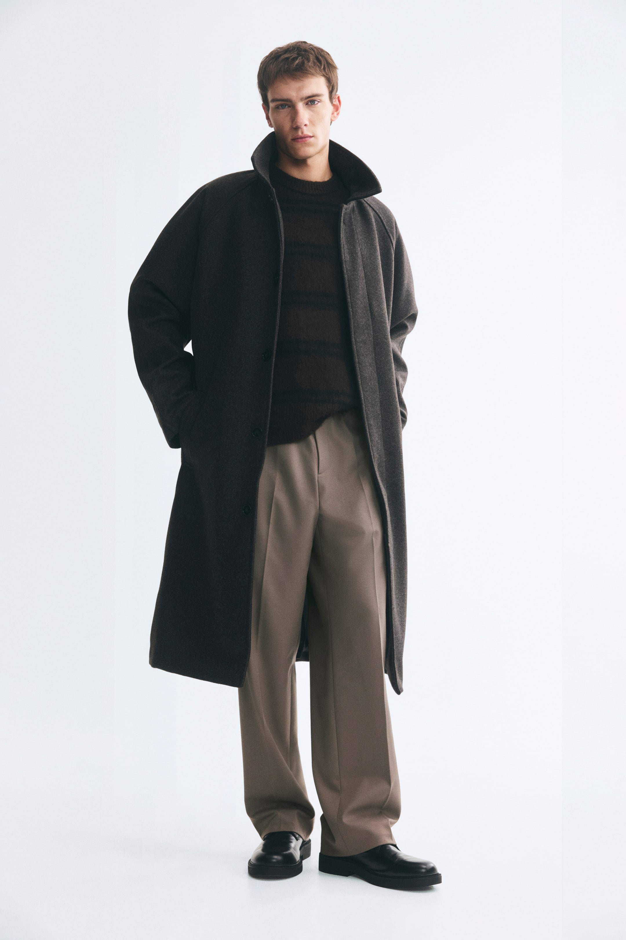 Größeres Bild ansehen: Maßgeschneiderte Jogger aus gewebtem Taupe-Stoff werden vom Model getragen und zeichnen sich durch eine entspannte Silhouette sowie markante Bügelfalten entlang der Vorderseite der Beine aus. Die Hose ist mit einem dunkel gestreiften Strickpullover, einem langen dunklen Mantel und schwarzen Lederschuhen kombiniert.