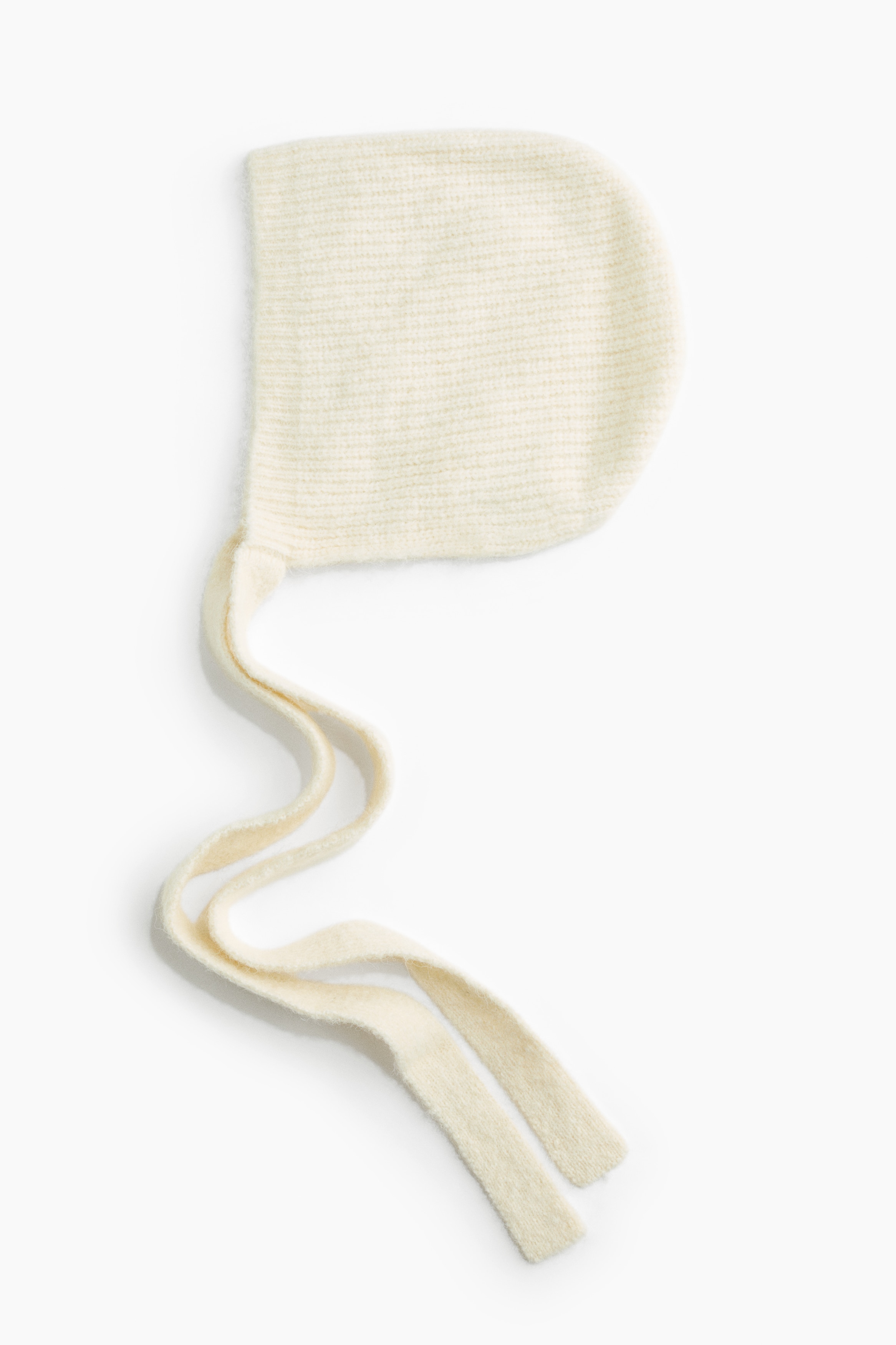 Een crèmekleurige balaclava van ribgebreide stof met een puntige bovenkant wordt gedragen door het model. De lange, platte gebreide bindlinten van de balaclava hangen losjes over een bijpassende jas met een gestructureerde, krullende pool.