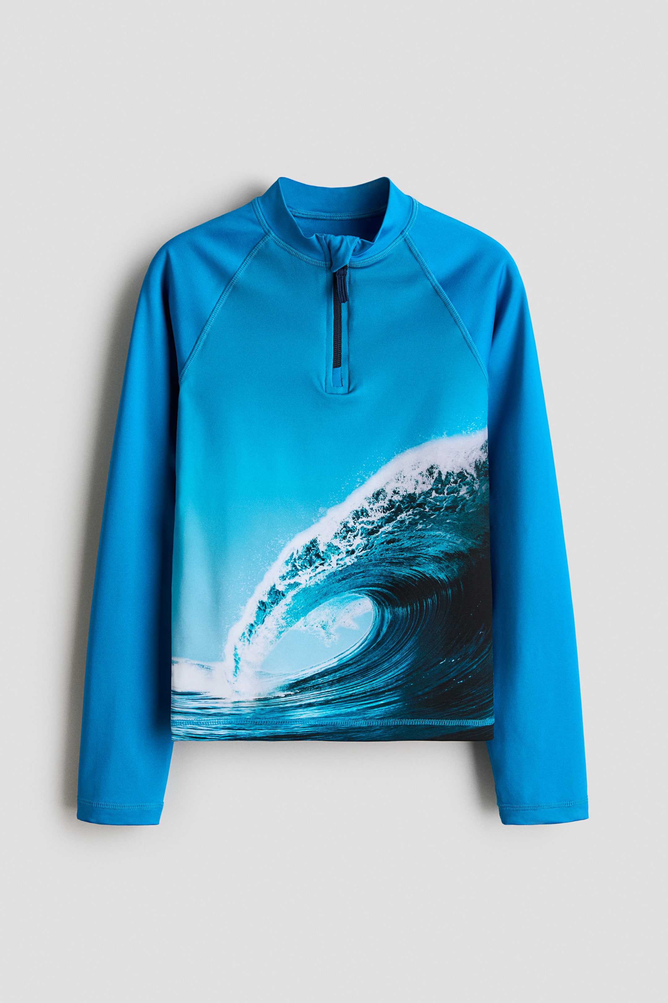 Visa större bild: En långärmad rash guard visas framifrån, med en ljusblå färg och en hög krage med en kort svart dragkedja. Ett stort, realistiskt våggrafik täcker den främre torson, och raglanärmar med synliga sömmar finns.