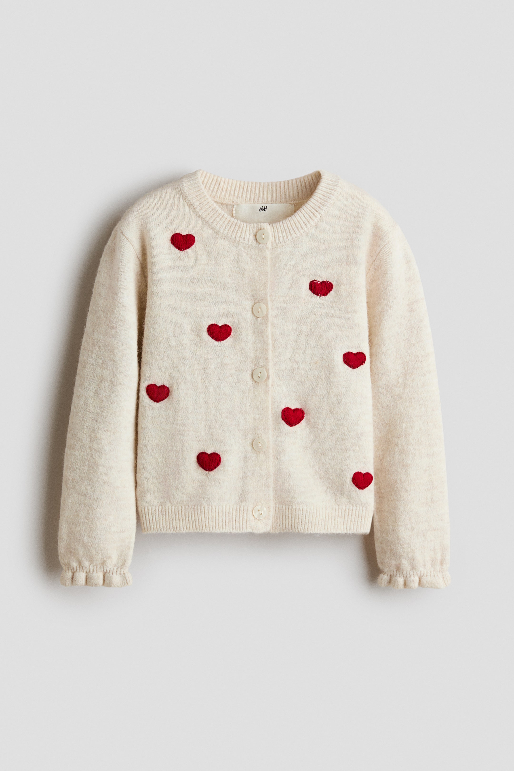 Agrandir l'image: Un cardigan de couleur crème en maille fine et douce est présenté de face, orné de motifs cœurs rouges brodés disséminés sur le devant. Il est conçu avec un col rond bordé de côtes, une fermeture boutonnée sur le devant, un bas côtelé et des manches longues avec poignets élastiqués à finition volantée.