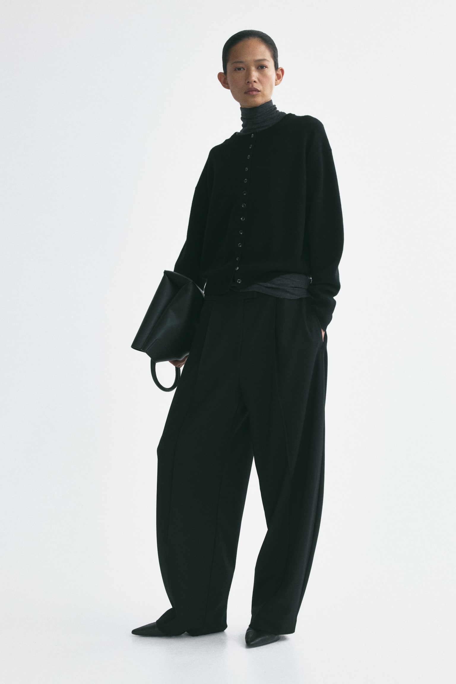 Un cardigan en laine noire tricotée, doté d’un col rond et d’une fermeture boutonnée sur le devant, est porté par le mannequin. Le cardigan présente des épaules tombantes et est superposé sur un haut à col roulé gris foncé, associé à un pantalon noir à jambes larges. - 1