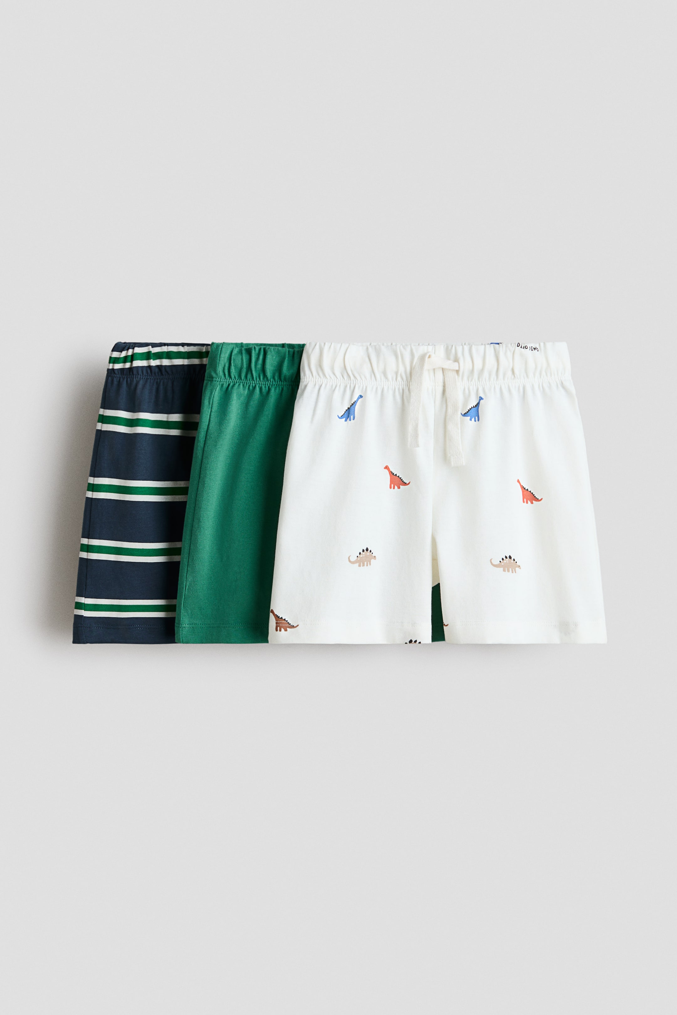 View larger image: Three pairs of children's shorts are shown from the front, arranged side-by-side. The left pair features navy blue with white horizontal stripes, the center pair is solid green, and the right pair is off-white with colorful dinosaur prints and an elastic waistband with a white drawstring tie.
