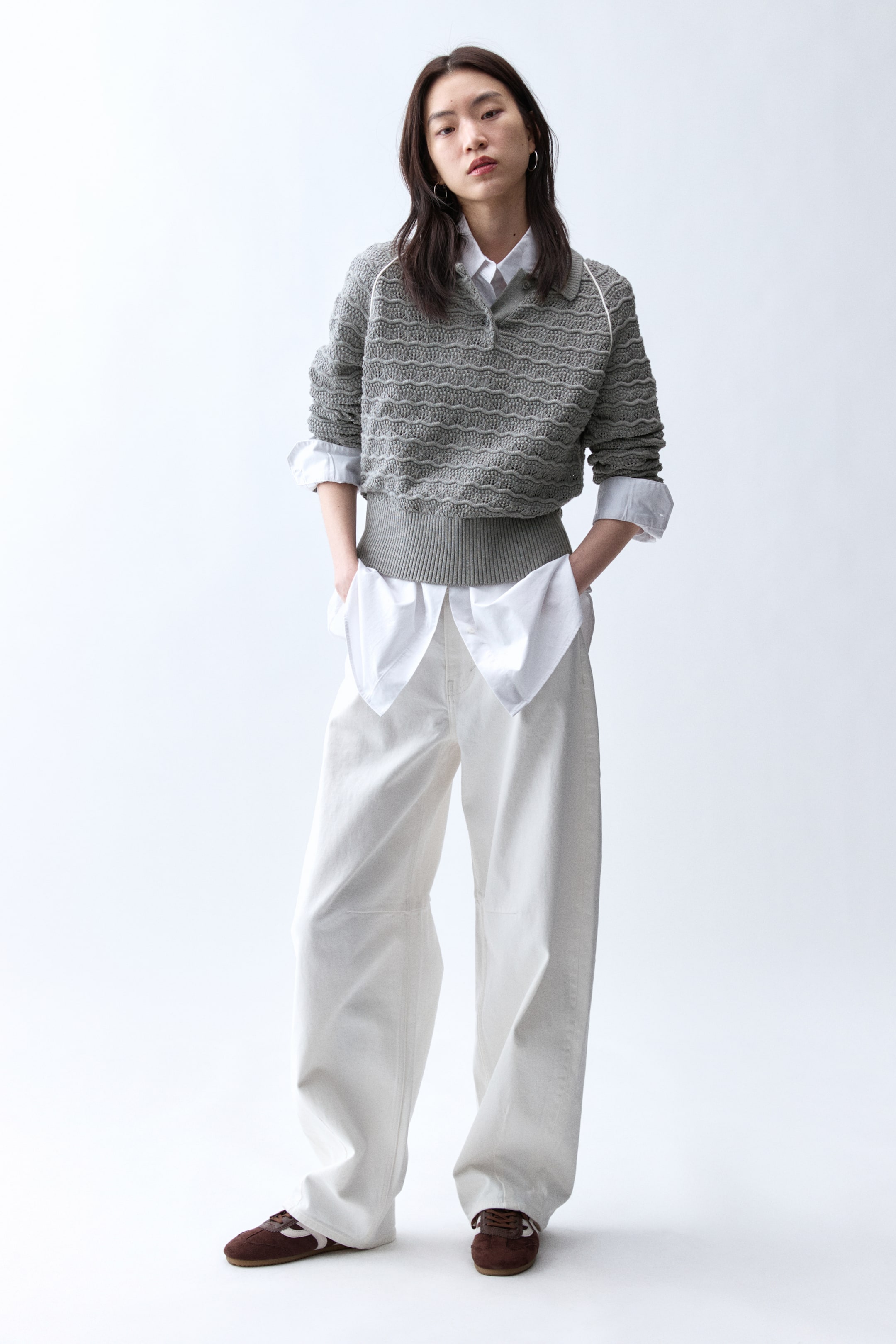 拡大画像を表示: グレーのニットポロセーターは、波状のテクスチャーと襟とボタンプラケットに沿った白いパイピングが特徴です。リブ編みの裾とカフスが施されており、長袖は白い襟付きシャツの上に押し上げてスタイリングされています。
