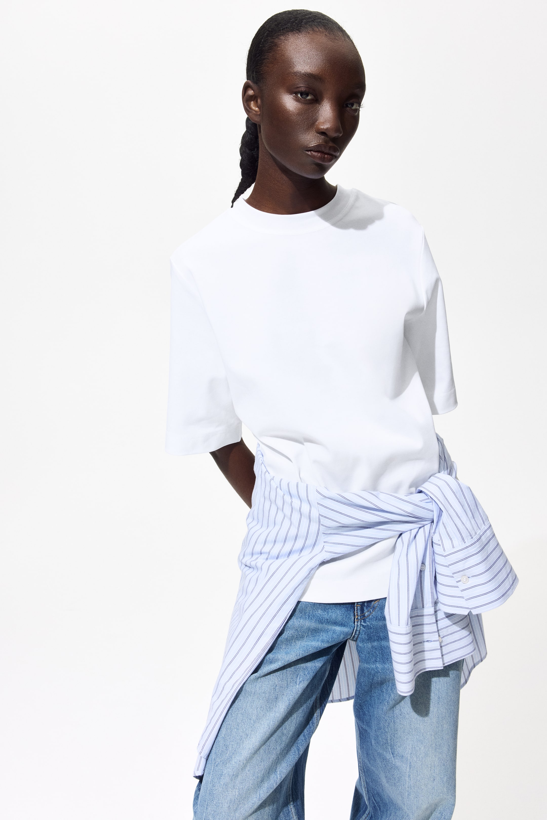 View larger image: The model is wearing a loose-fit white T-shirt, complete with a ribbed trim around the neckline and subtly dropped shoulders, paired with a blue and white striped shirt tied around the waist.