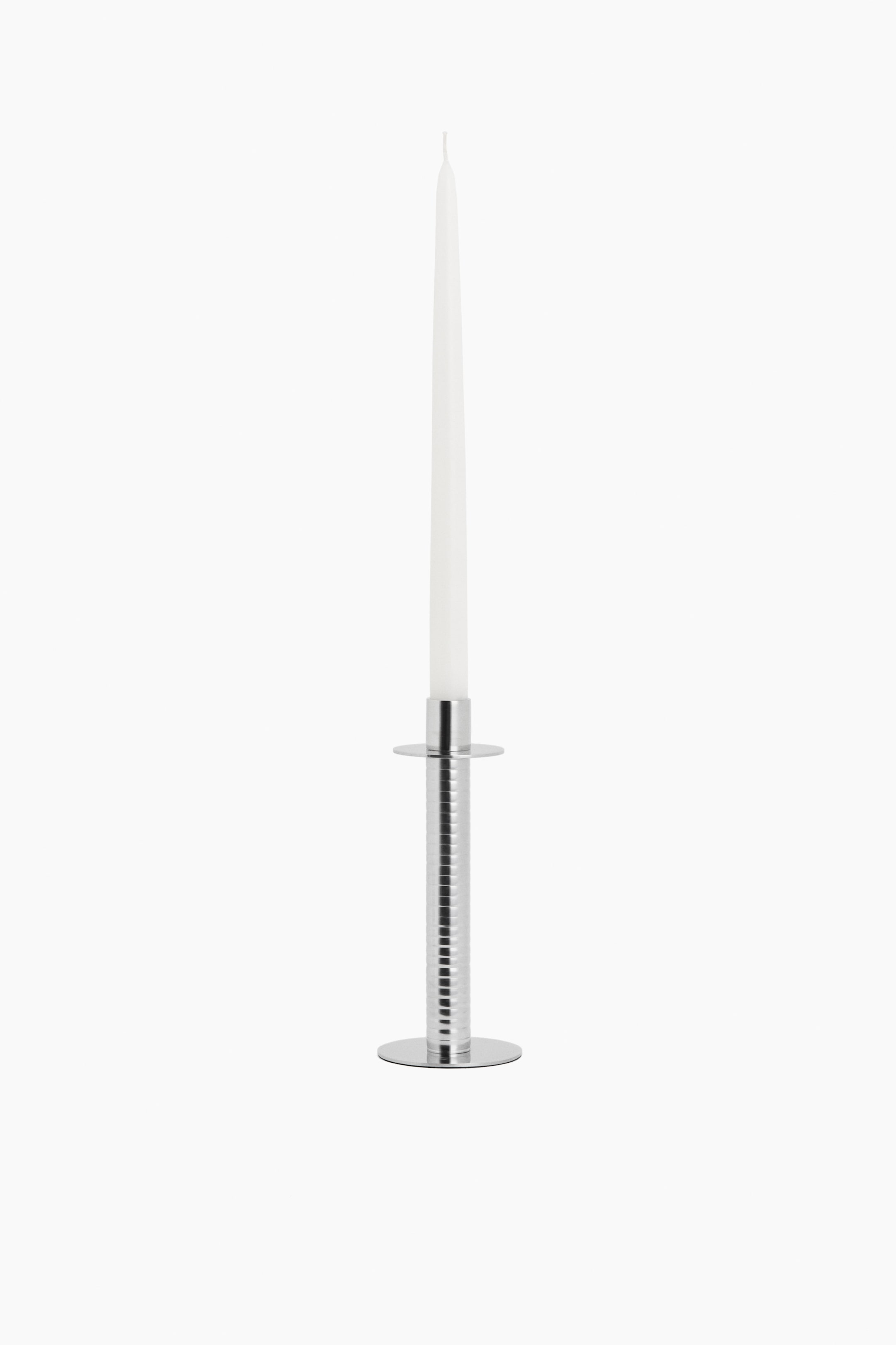 View larger image: A silver-toned metal candlestick holds a white taper candle. The candlestick showcases a ribbed cylindrical stem, a flat circular base, and a circular drip tray.