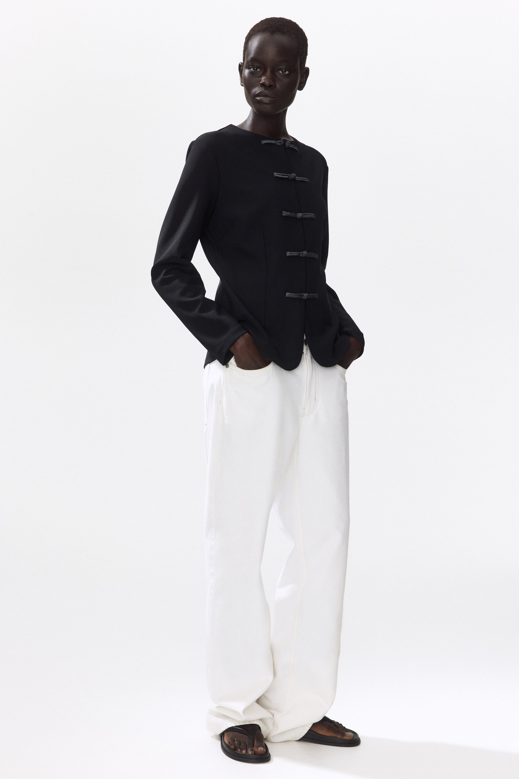 View larger image: The model is wearing a tailored black jacket featuring a round neckline and knotted buttons running down the front. Waist darts create a cinched silhouette. Paired with the jacket are white wide-leg pants.