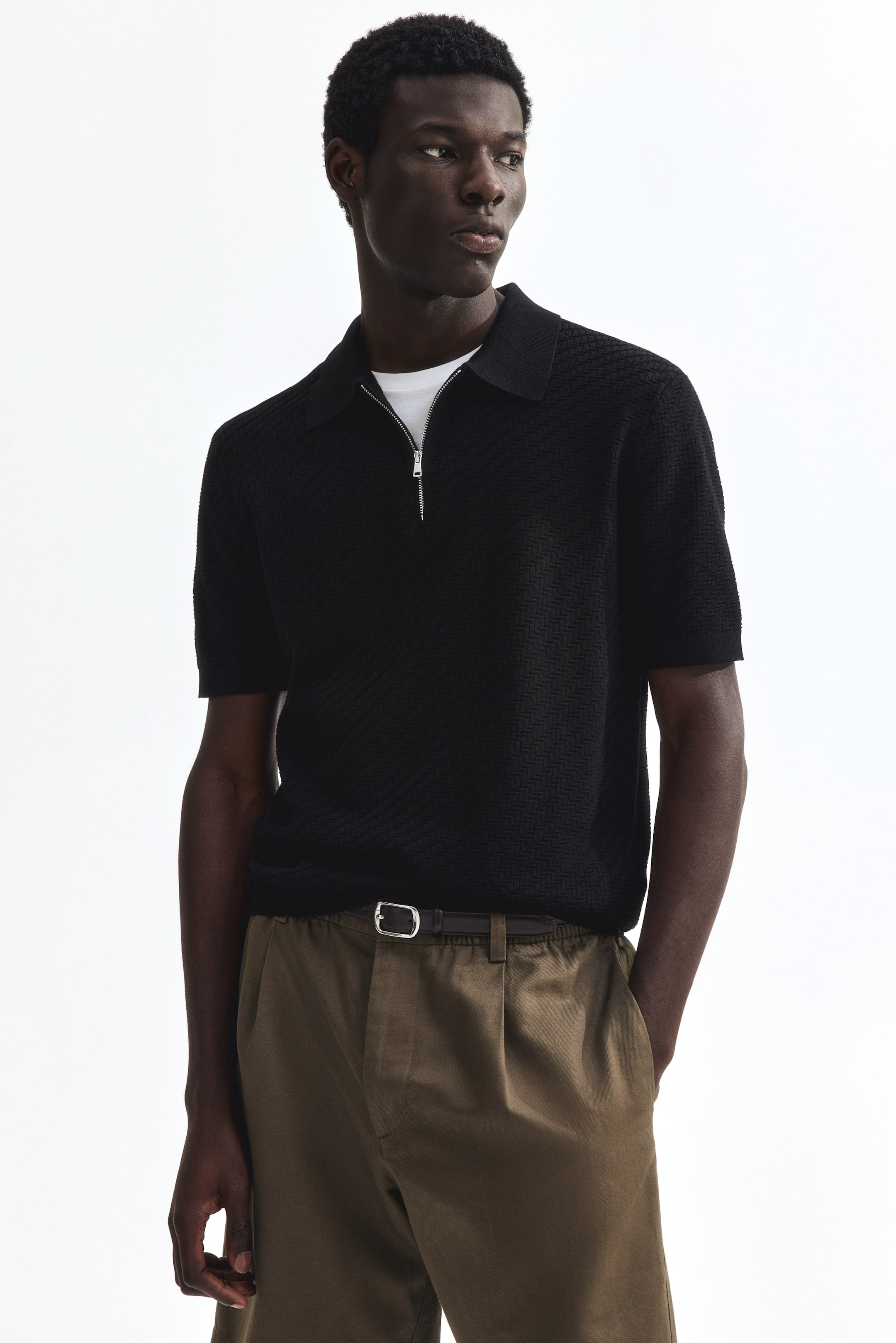 View larger image: The black knit polo shirt boasts a textured geometric pattern, short sleeves, and a standard collar. It features a silver half-zip placket, with a white crew neck t-shirt subtly visible underneath.