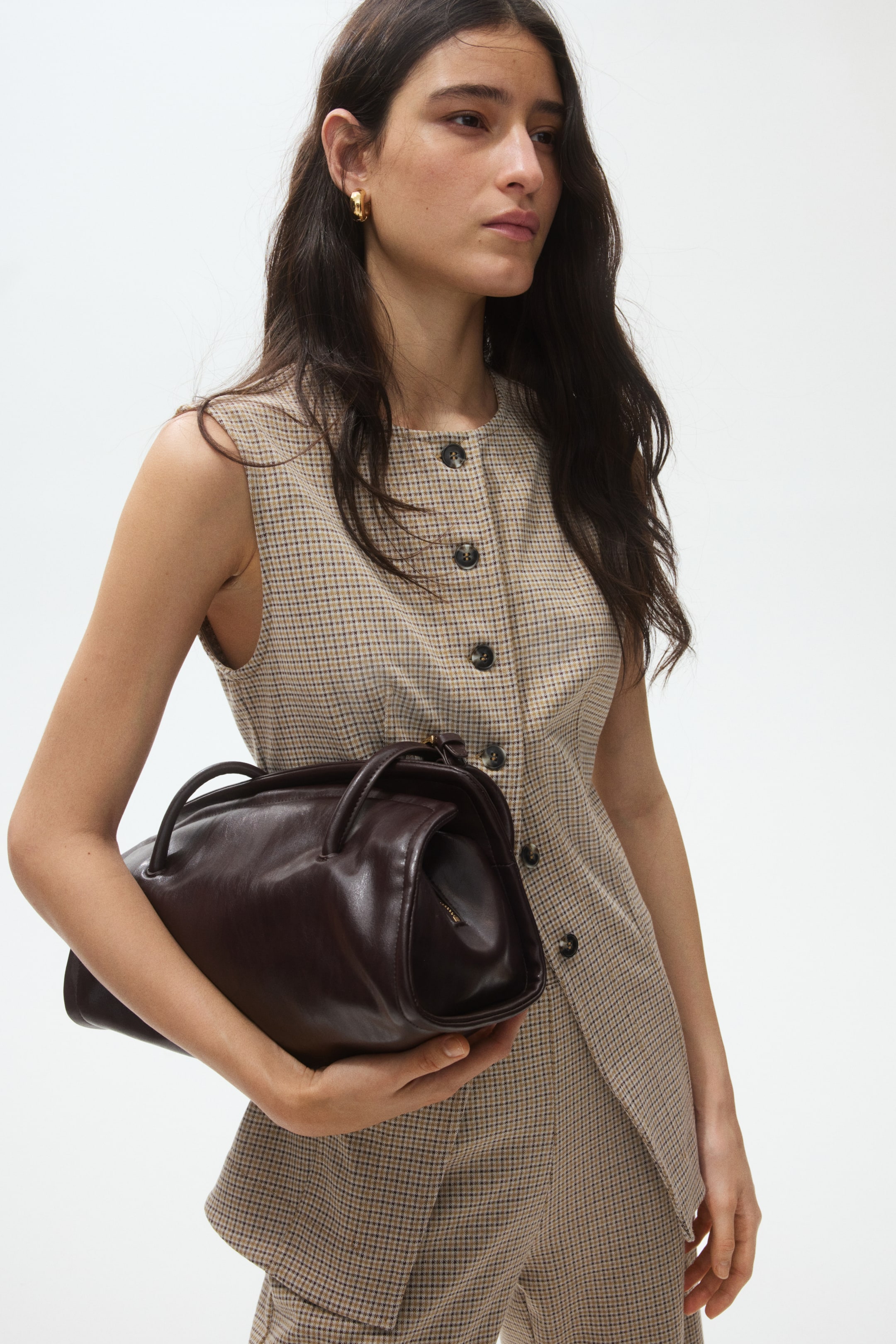 View larger image: A dark brown leather handbag is being held horizontally, highlighting its structured, elongated shape with a rounded top, two short top handles, and a visible zip closure running along its upper edge.