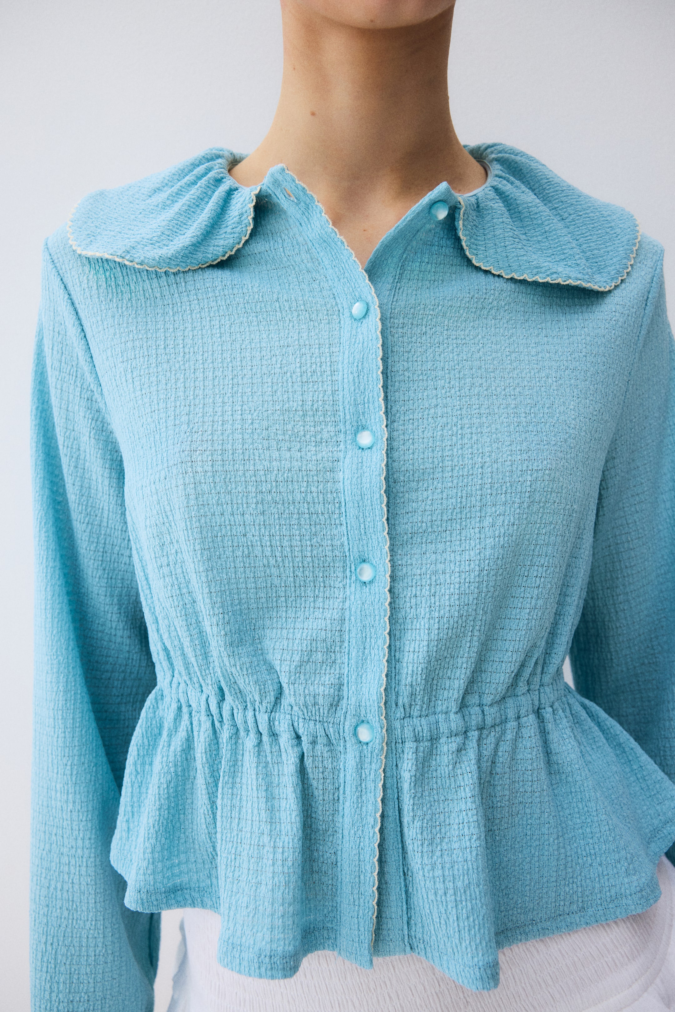 View larger image: "A light blue textured peplum top featuring a rounded, gathered collar with a cream scalloped edge. It showcases a button-front placket with matching cream trim and an elasticized waist forming a flared peplum hem."