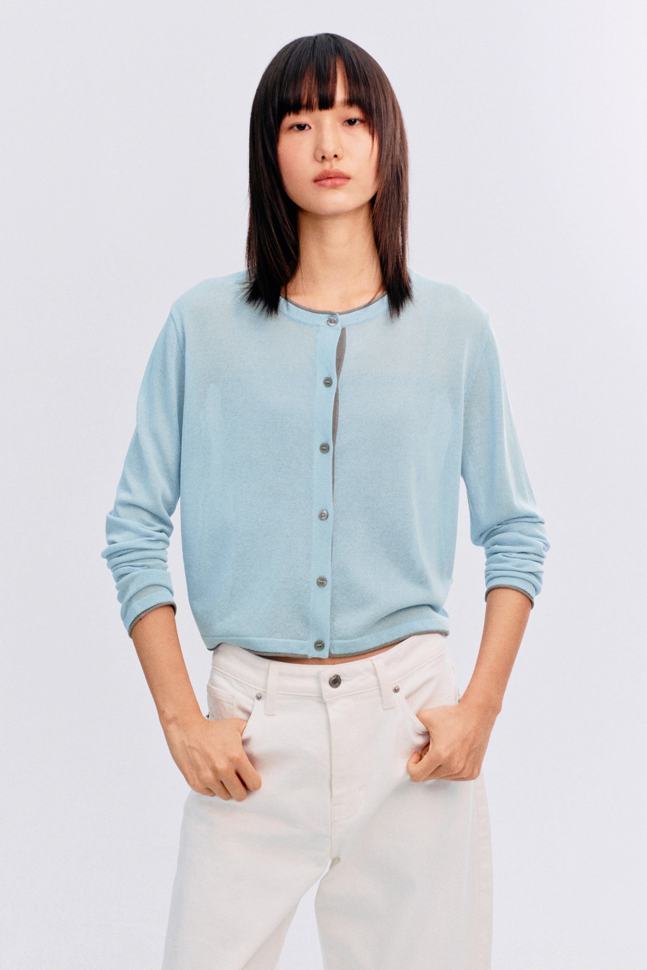拡大画像を表示: ライトブルーのカーディガンは、ラウンドネックラインと小さなグレーのボタンが並ぶボタンフロントクロージャーを備えています。長袖、クロップドヘム、前立てと袖口に沿ったグレーのトリムが特徴です。