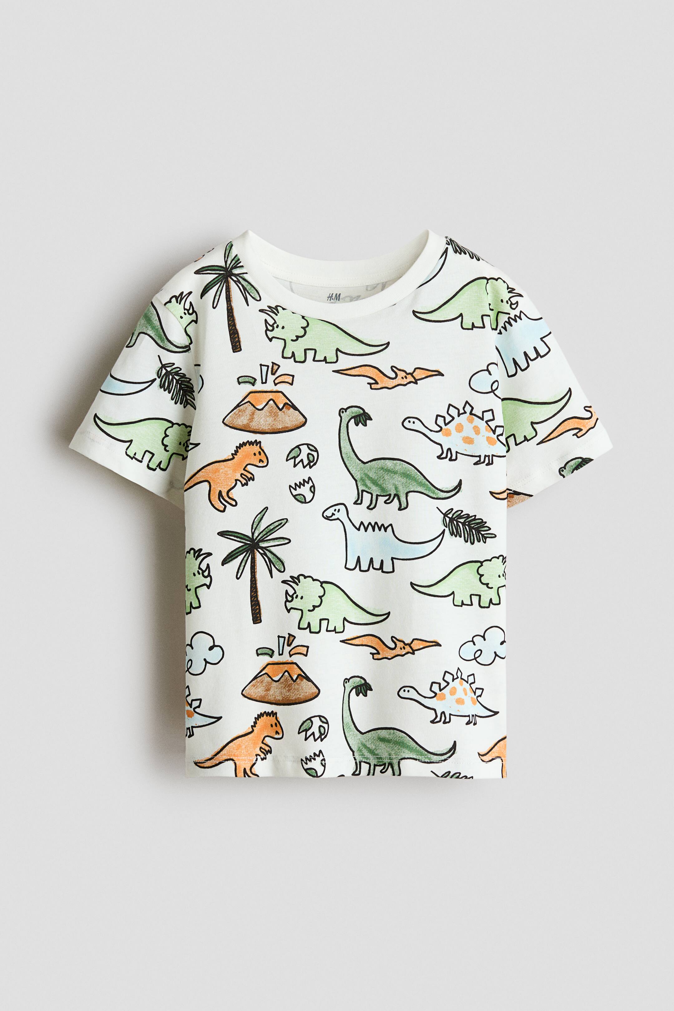 View larger image: A white, short-sleeved T-shirt shown from the front, featuring an all-over cartoon print of assorted dinosaurs, palm trees, and erupting volcanoes in muted green, orange, blue, and brown tones. The T-shirt has a ribbed crew neck and a straight hem.