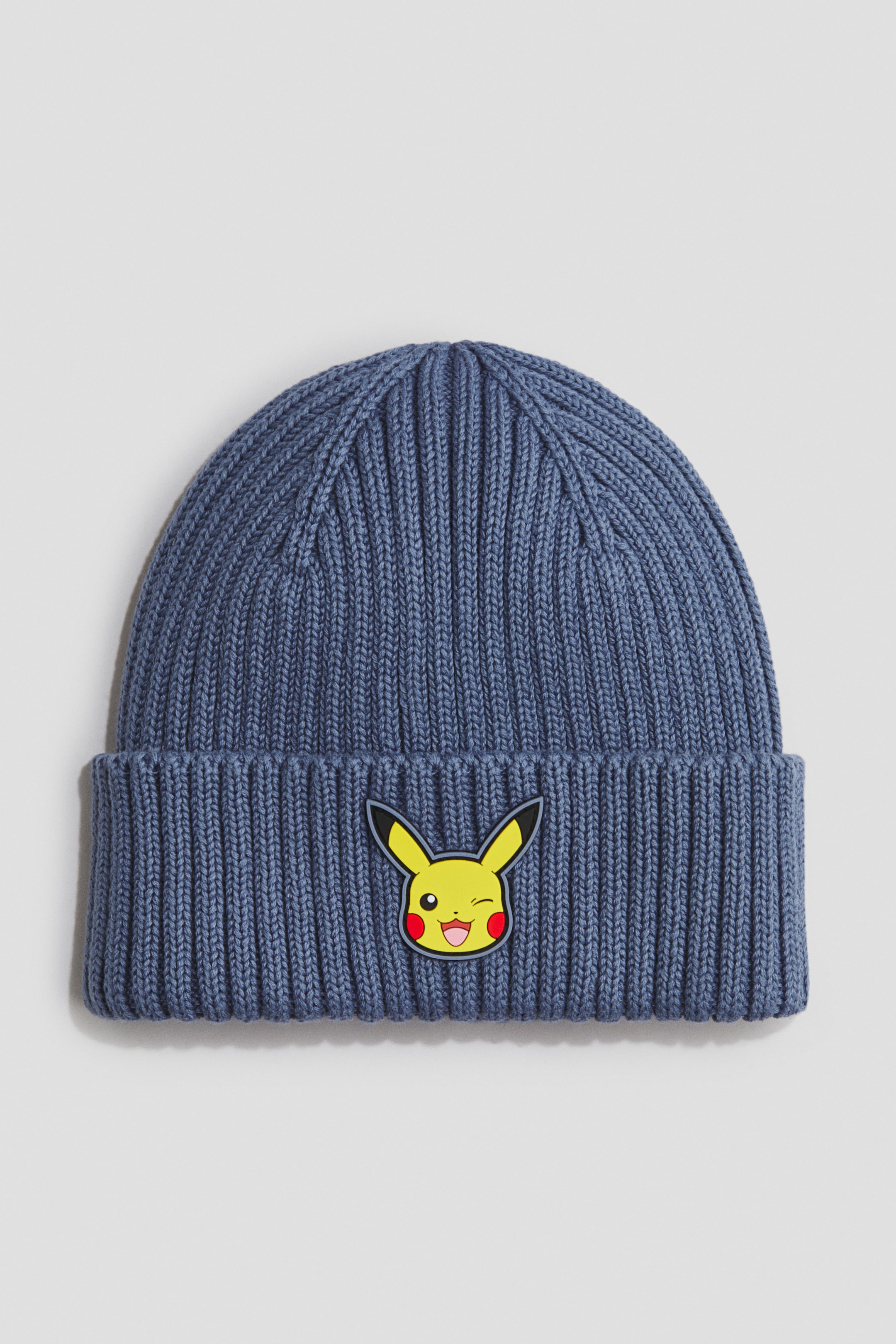 Voir une image plus grande: Un bonnet tricoté côtelé bleu affiche un écusson de Pikachu clin d’œil jaune sur son bord retroussé.