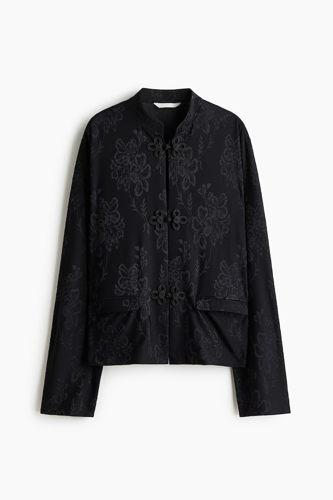 A black, relaxed-fit jacket in a tonal floral jacquard pattern is hanging front-facing, showcasing a band collar, dropped shoulders, and three decorative knot buttons down the centre. The jacket features long sleeves and jetted front pockets with flaps. - 1