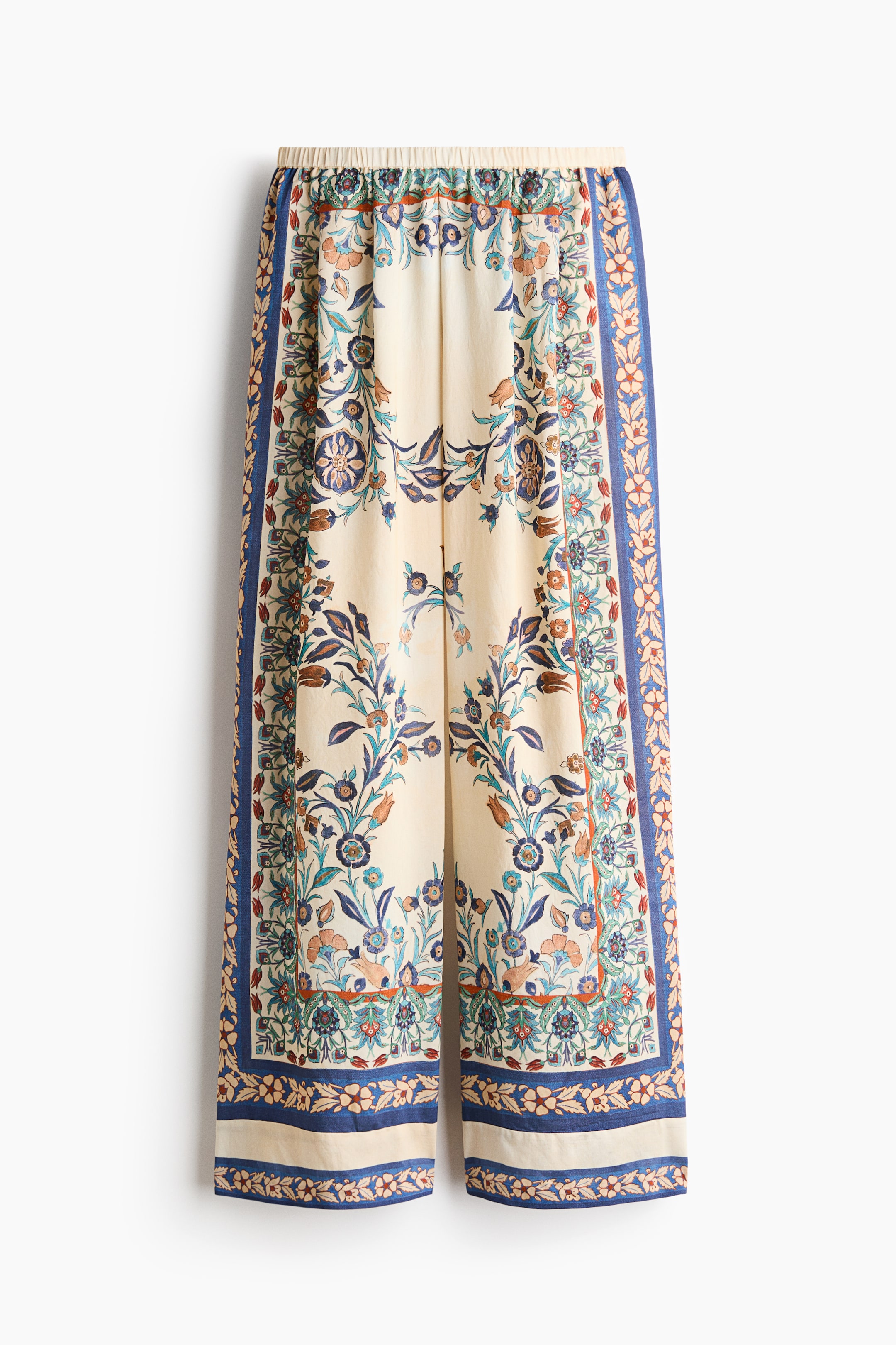 View larger image: Trousers showcase an intricate floral and foliage design in hues of blue, teal, orange, and brown on an off-white background, viewed from the front. They feature an elasticated waistband and wide legs, with decorative vertical borders along the outer seams and horizontal borders at the hems.