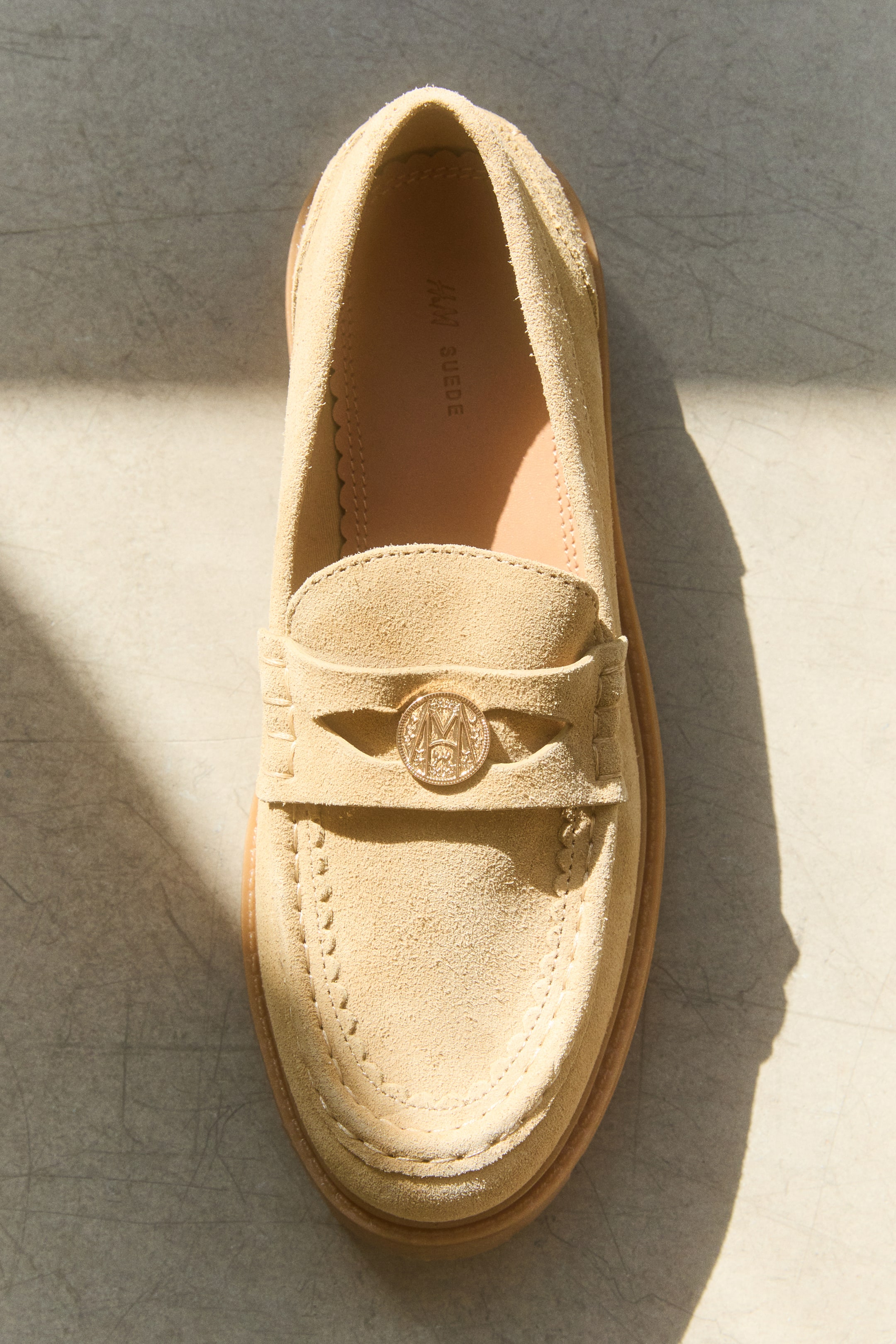 View larger image: An overhead view showcases a beige split-suede loafer featuring a stitched moccasin seam and a classic penny keeper strap across the front. A decorative, gold-coloured metal coin detail is nestled in the centre of the strap, while a chunky light-brown sole frames the shoe.