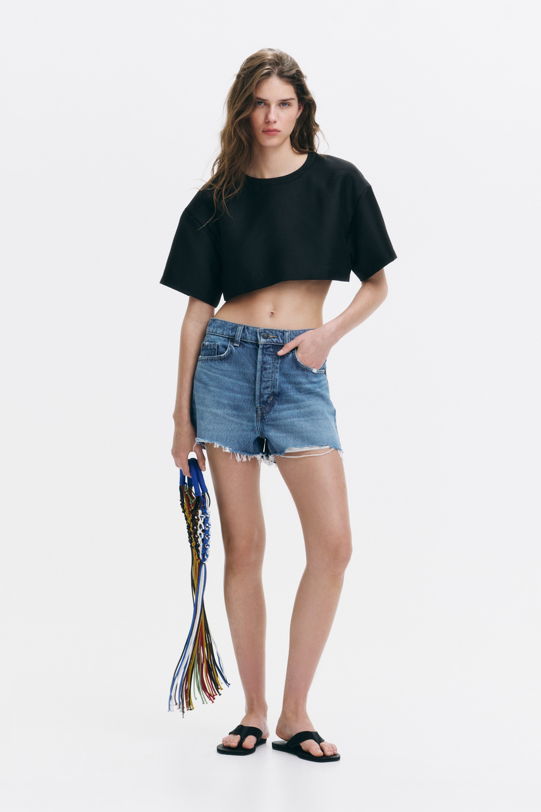Se større billede: 5-lomme shorts i stift bomuldsdenim med høj talje og knaplukning og rå kanter bæres af modellen. Modellen står med den ene hånd i en forreste lomme på shortsene, mens hun også har en sort cropped top og sorte sandaler på.
