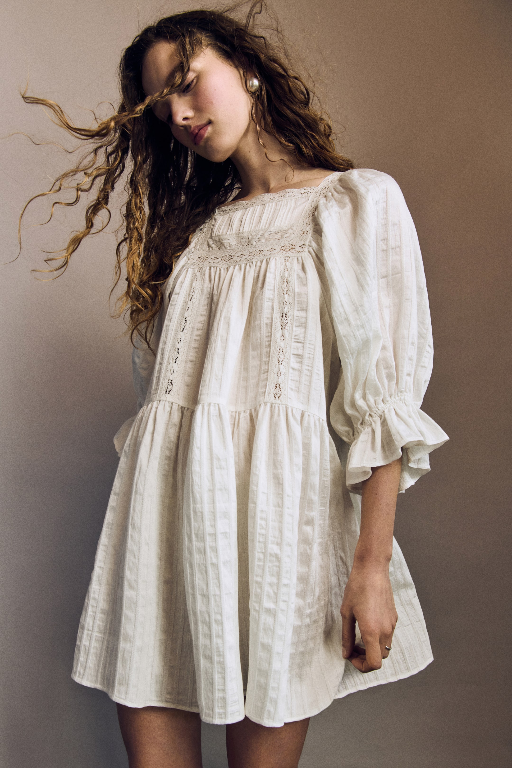 Ver imagem maior: Um vestido curto e de corte largo, confeccionado em tecido texturizado e adornado com inserções de renda, é usado pela modelo. Foi desenhado com um decote quadrado com acabamento em renda, um canhão estreito com pregas na frente, mangas volumosas de 3/4 de comprimento com elástico nos pulsos e punhos em evasé, uma costura franzida na cintura e uma saia suavemente evasé.