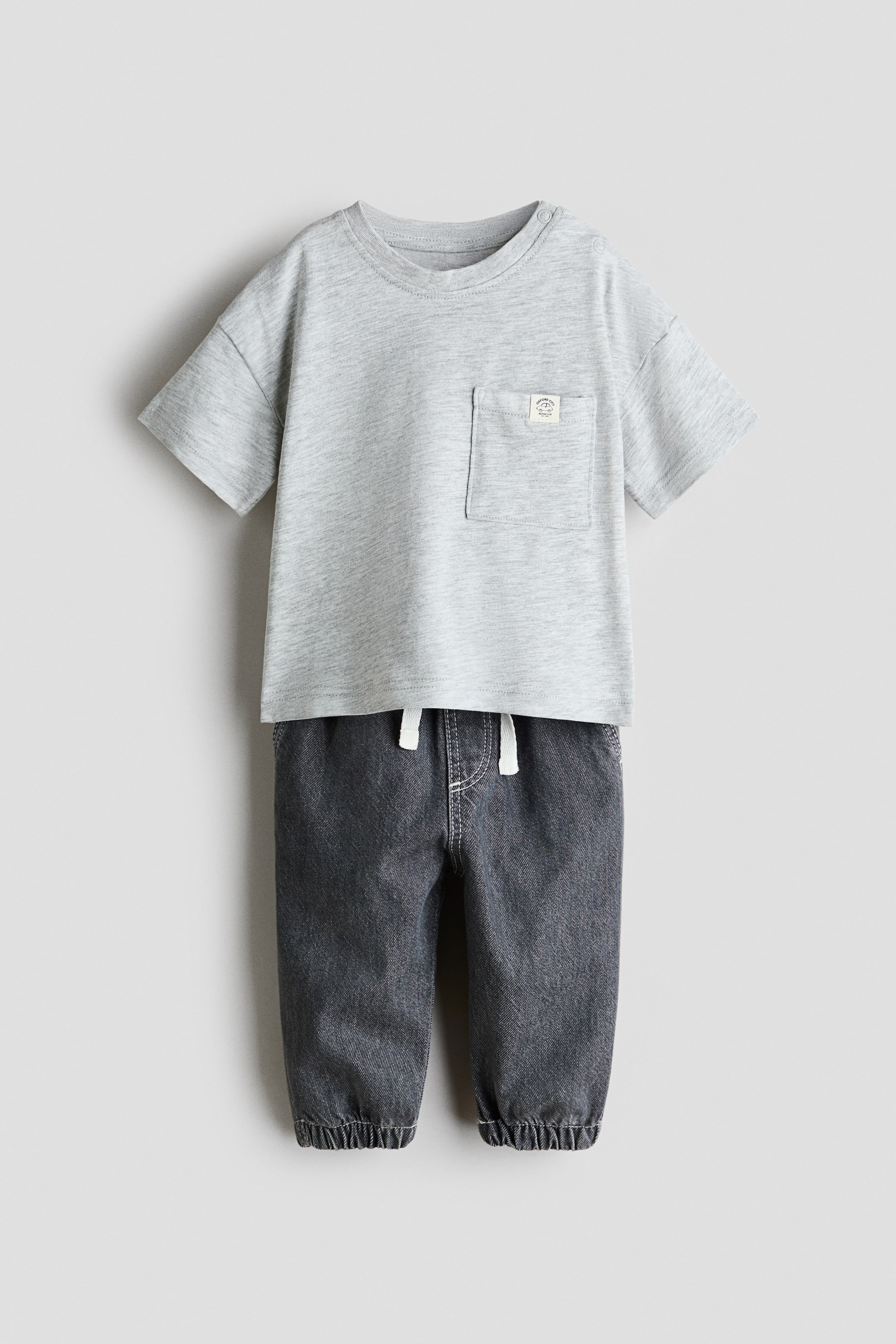 Visa större bild: En ljusgrå melerad kortärmad t-shirt med rund hals och en bröstficka på vänster sida visas, med tryckknappar vid axlarna. Den matchas med mörkgrå denim-joggers med resår i midjan och vita dragsnören samt resår i bensluten; båda plagg presenteras framifrån.
